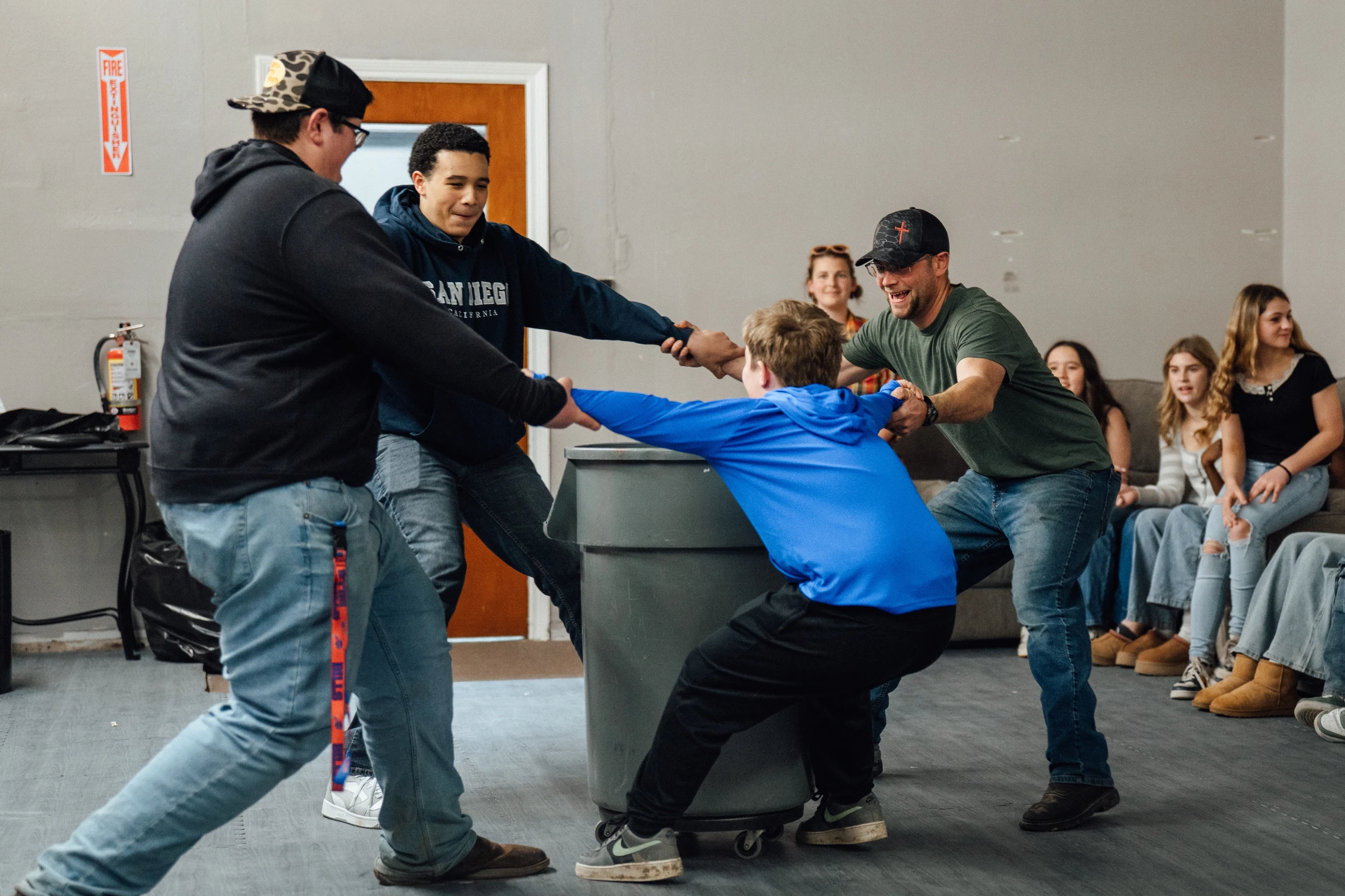 A group of smiling men and boys play a tug-of-war game, pulling on a boy in a blue shirt centered around a large grey trash can, as others watch.