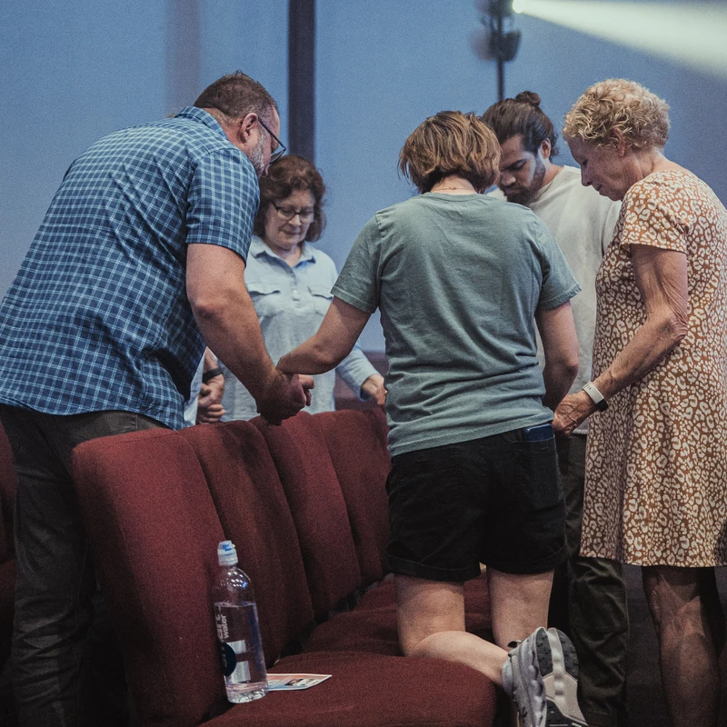 A diverse group of people stands in a circle holding hands and bowing their heads in prayer. One woman kneels on a red pew in front of a wooden cross.
