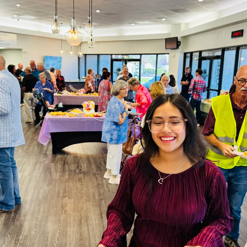 A smiling young woman in a magenta blouse is in the foreground of a busy indoor gathering, with many people socializing and eating in the background.