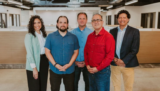 A group of five diverse colleagues, one woman and four men, standing together and smiling for a photo in a modern, open-plan office space.
