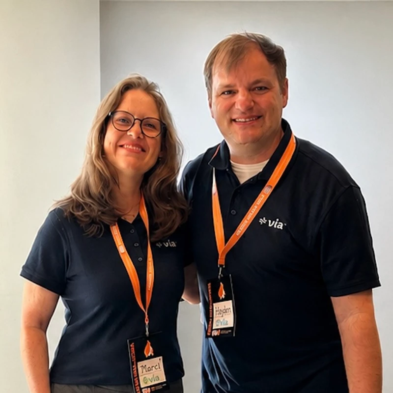 A woman with glasses and a man stand side-by-side, both smiling and wearing matching dark blue polo shirts and orange lanyards with conference badges.