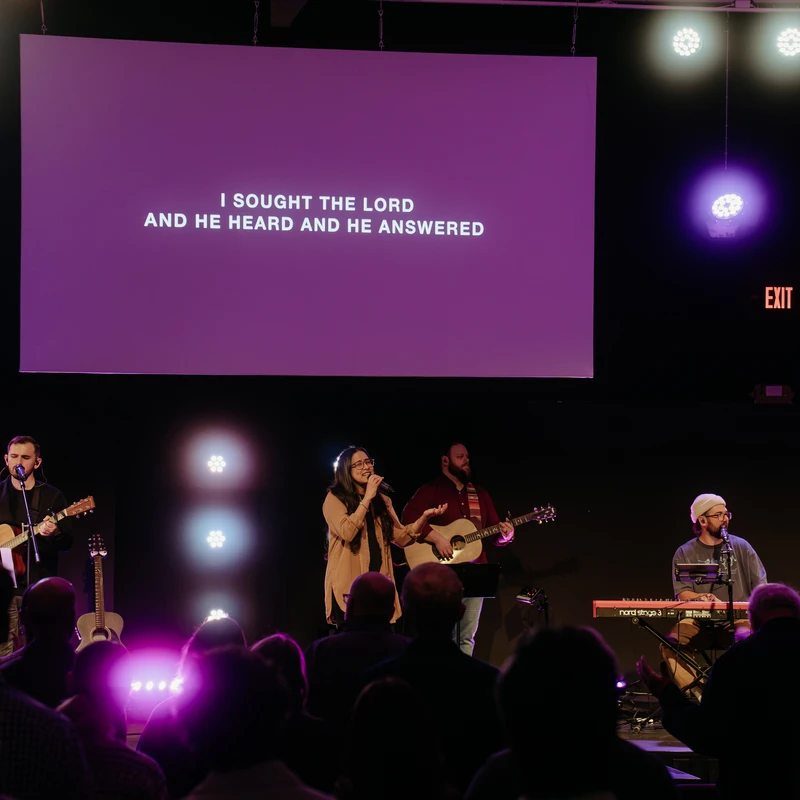 A band performs on a dimly lit stage, with a large purple screen behind them displaying lyrics. The audience watches from the foreground.