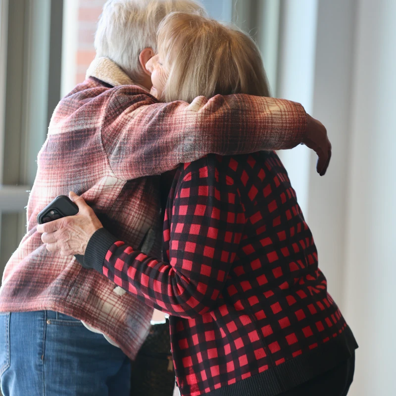 Two older women in plaid jackets share a warm hug while standing indoors near a window. One woman has white hair and the other has blonde hair.