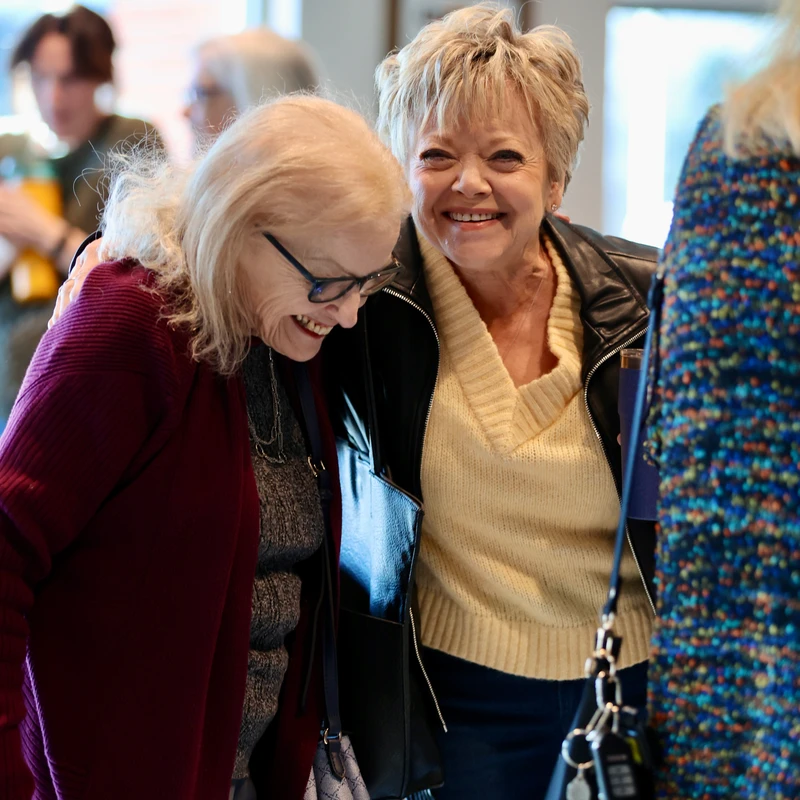Two senior women are smiling and laughing together. One woman in a black leather jacket has her arm around her friend, who is wearing a maroon cardigan.