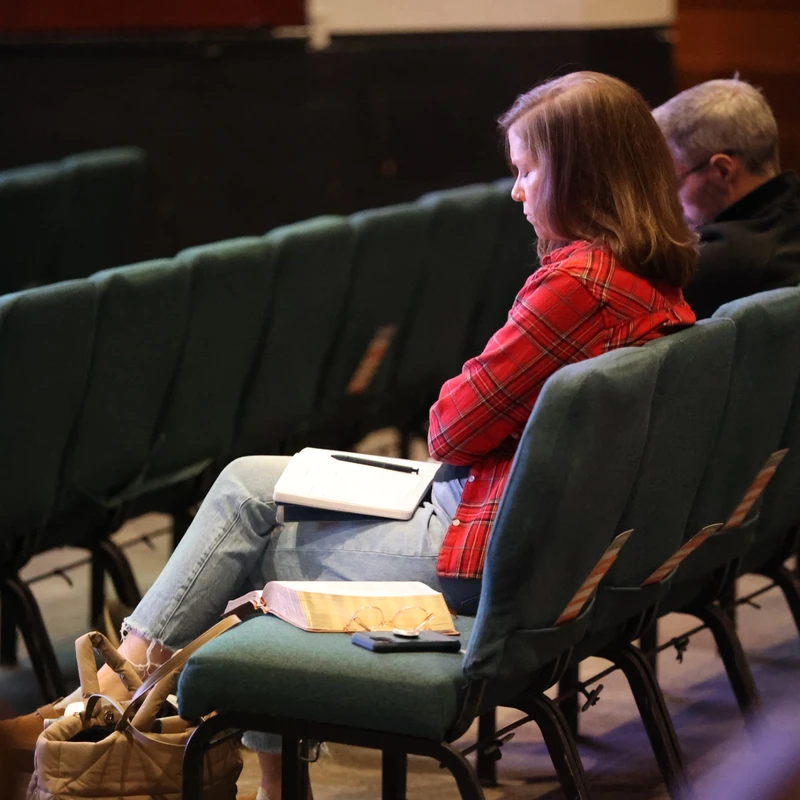 A woman in a red plaid shirt sits in a row of green chairs, looking down to study. A notebook is on her lap and an open Bible is on the seat next to her.