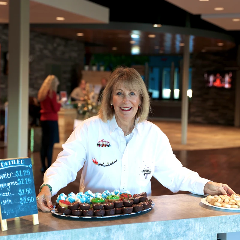 A smiling woman in a white Christmas-themed shirt stands behind a counter, offering a platter of small brownies and colorfully frosted desserts.