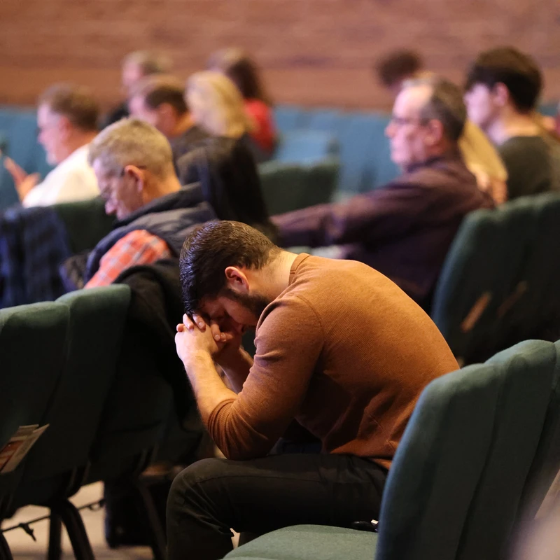 A man in a brown shirt sits in a theater-style chair, head bowed and hands clasped to his face in prayer. Other people are seated in rows in the background.