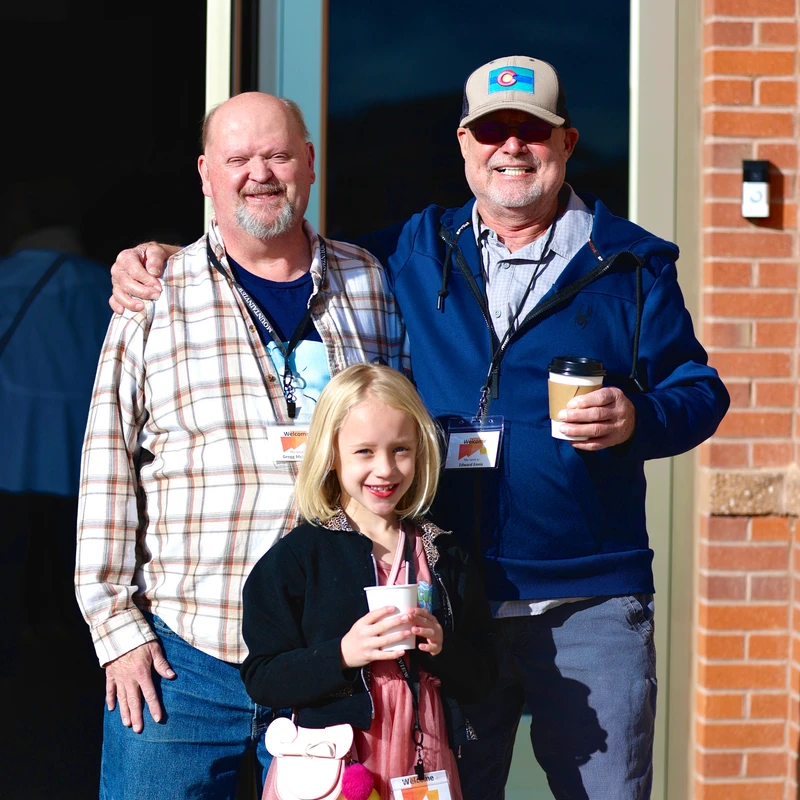 A young blonde girl in a pink dress stands smiling in front of two smiling men. They are outdoors by a brick building and wearing "Welcome" name tags.