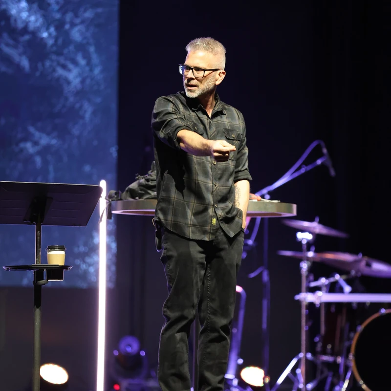 A man with short gray hair and glasses, wearing a plaid shirt and dark pants, stands on a dimly lit stage, speaking and gesturing with one hand.
