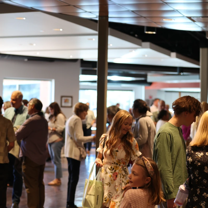 A large group of casually dressed people of various ages gathered in a modern, well-lit hall, standing in small groups and talking amongst themselves.