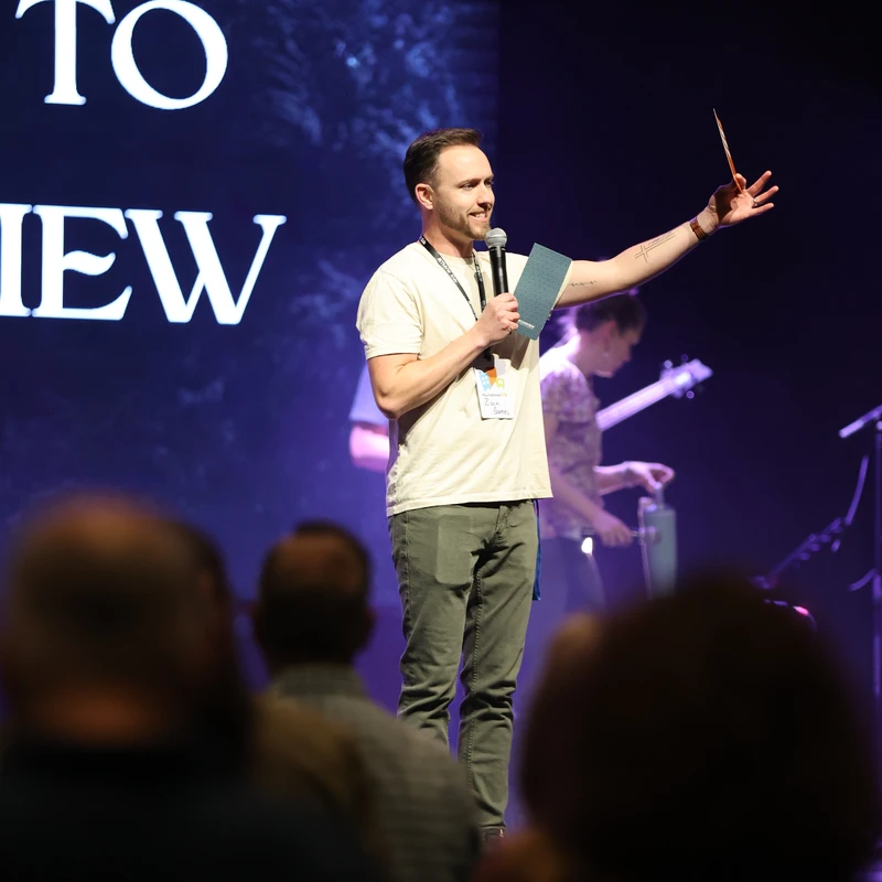 A smiling man stands on a dimly lit stage, speaking into a microphone. He holds a card and gestures towards the audience, who are visible in the foreground.