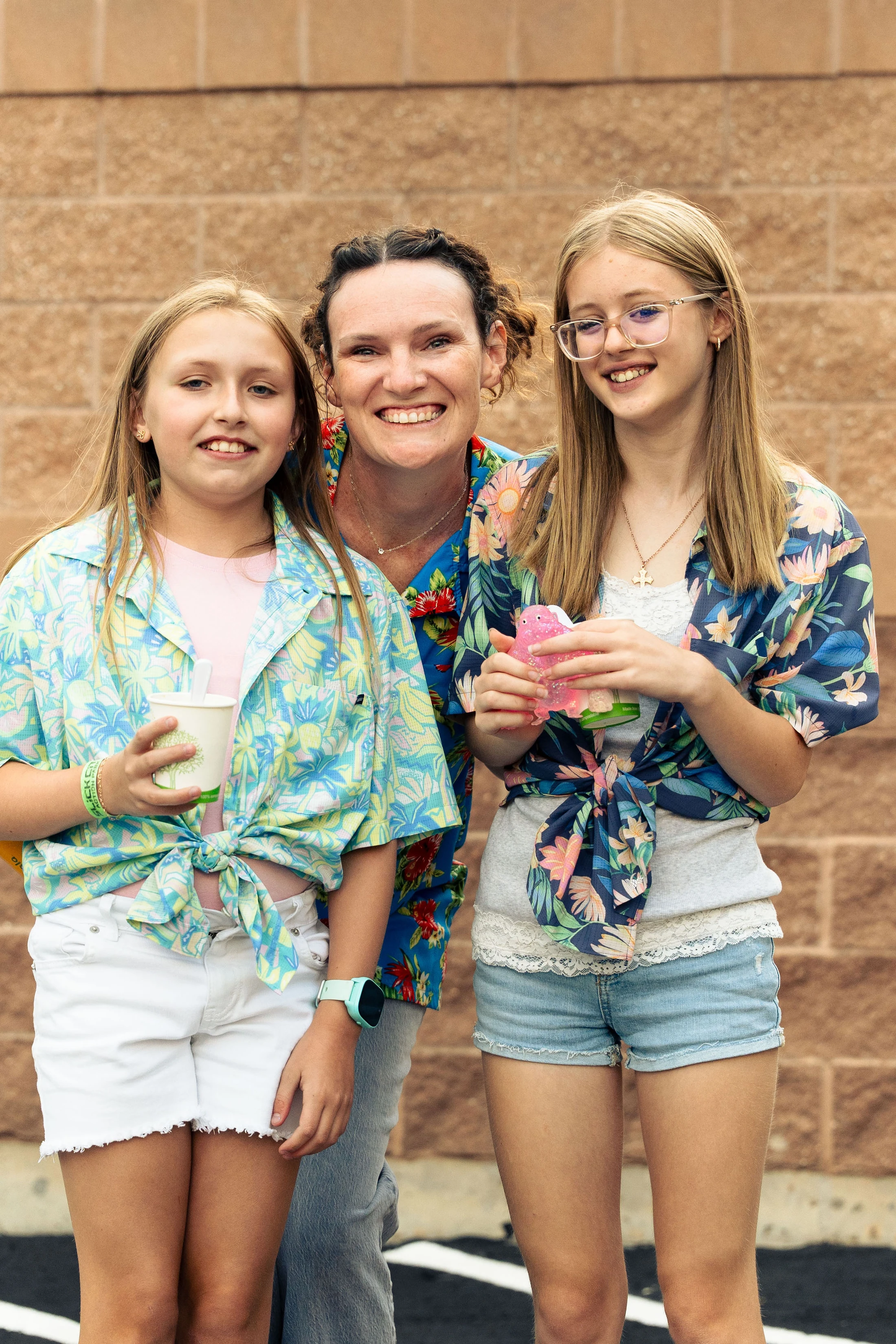 A smiling woman stands between two young girls against a brick wall. They are all wearing colorful, tropical-patterned shirts and smiling for the camera.