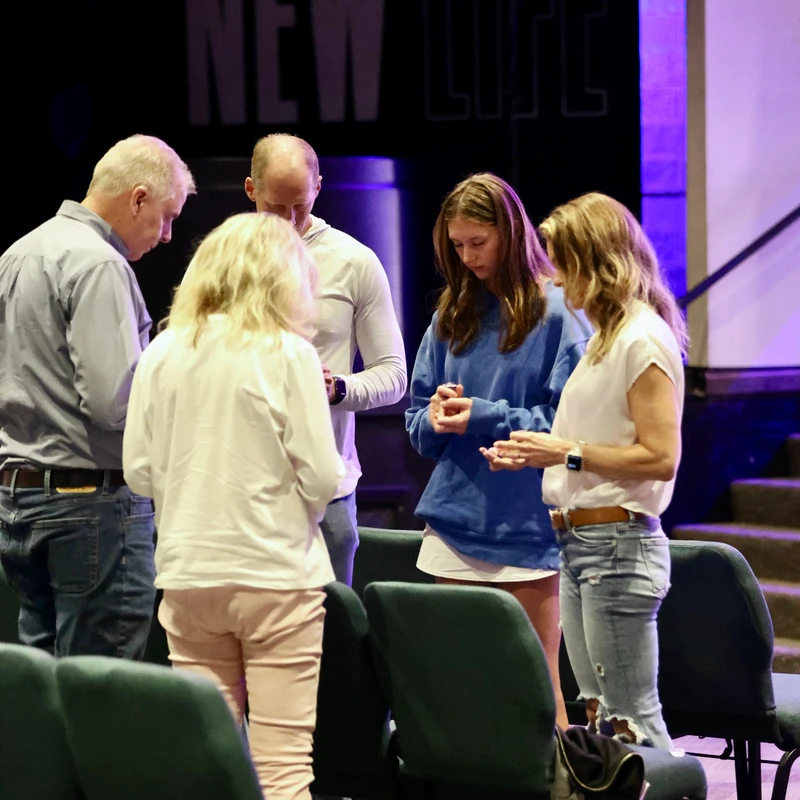 A group of five people, a mix of adults and a young girl, stand in a circle with their heads bowed in prayer inside a church with a purple-lit stage.