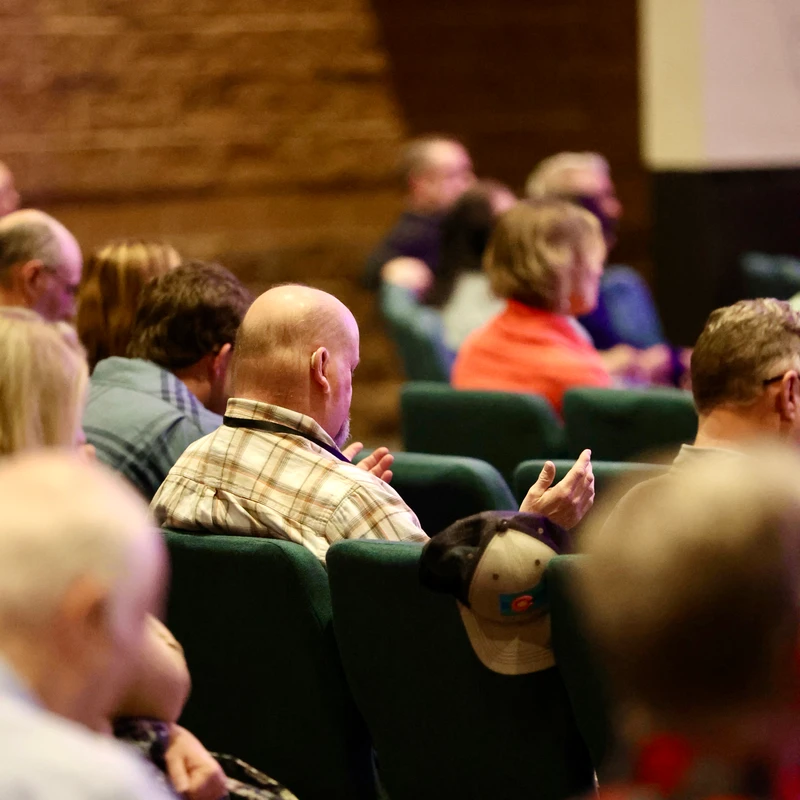 A view from behind of people sitting in an auditorium. The focus is on a bald man in a plaid shirt listening attentively.