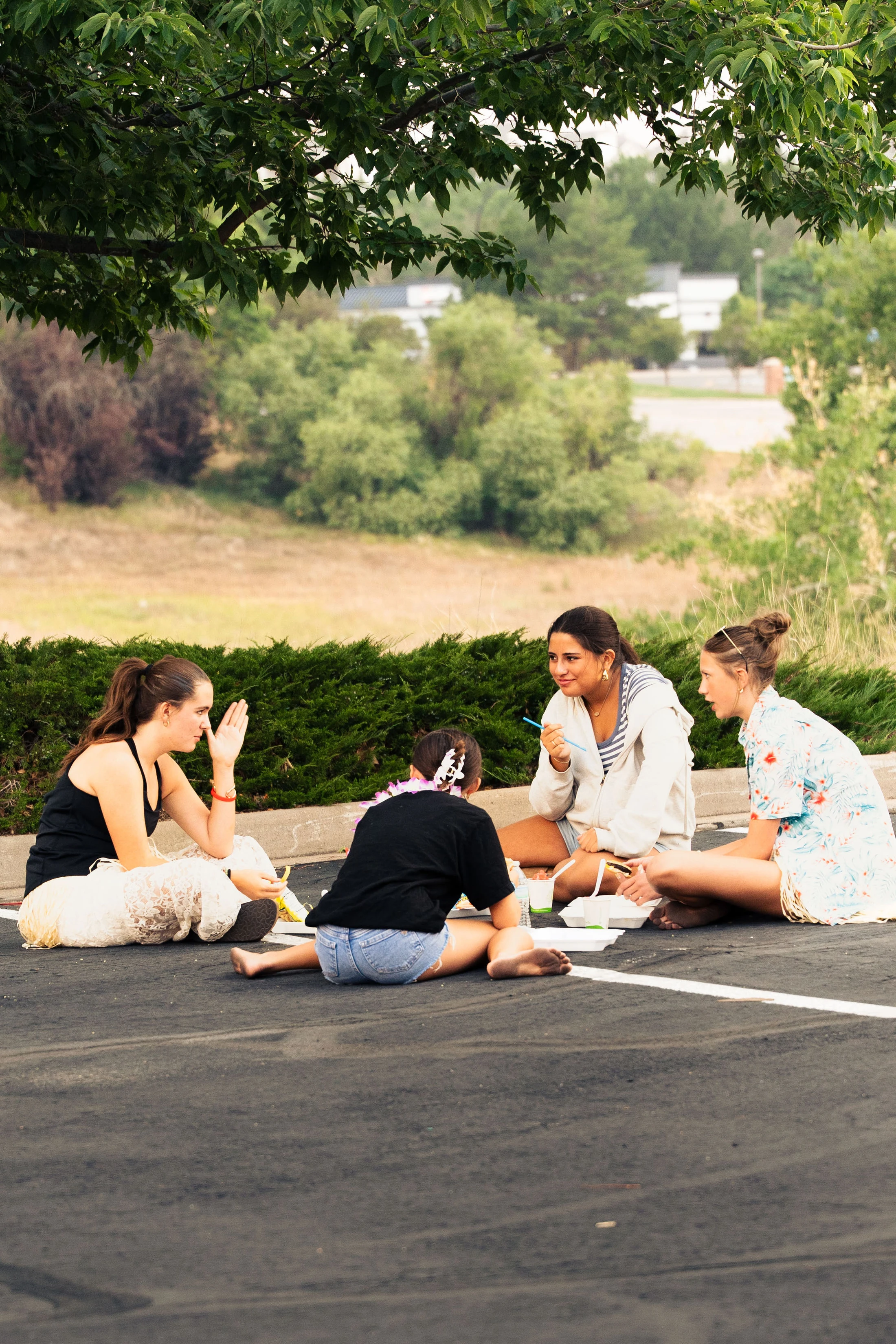 Four young women sit in a circle on the asphalt of a parking lot, eating and chatting together under the shade of a large green tree.