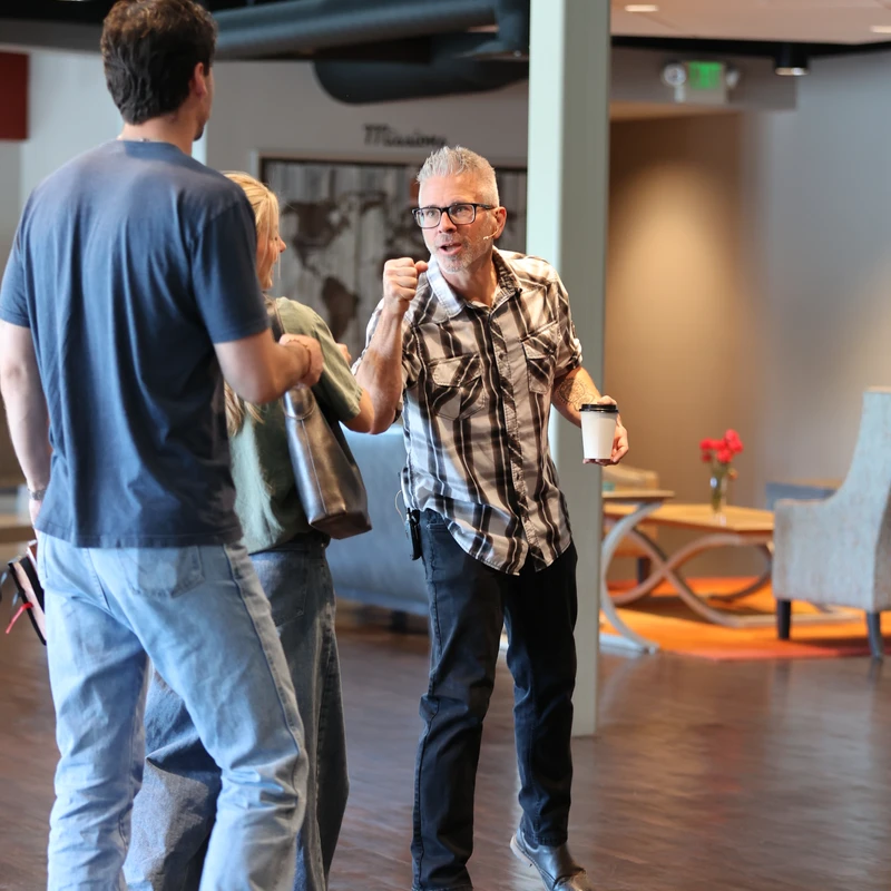 A man with grey hair and glasses gestures with his fist as he speaks animatedly to a man and woman in a modern building lobby while holding a coffee cup.