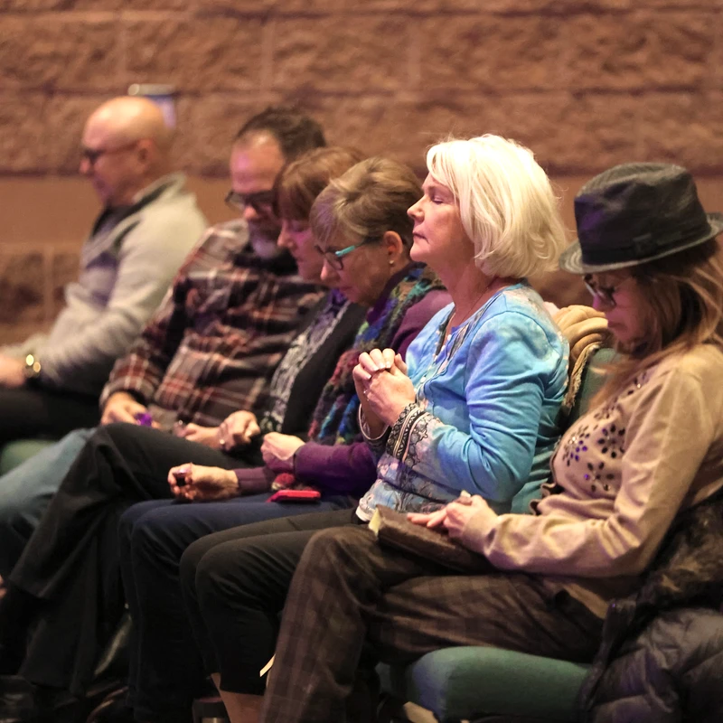 A row of men and women sitting in chairs against a brick wall. A woman in a blue shirt has her hands clasped as if in prayer, her eyes closed.