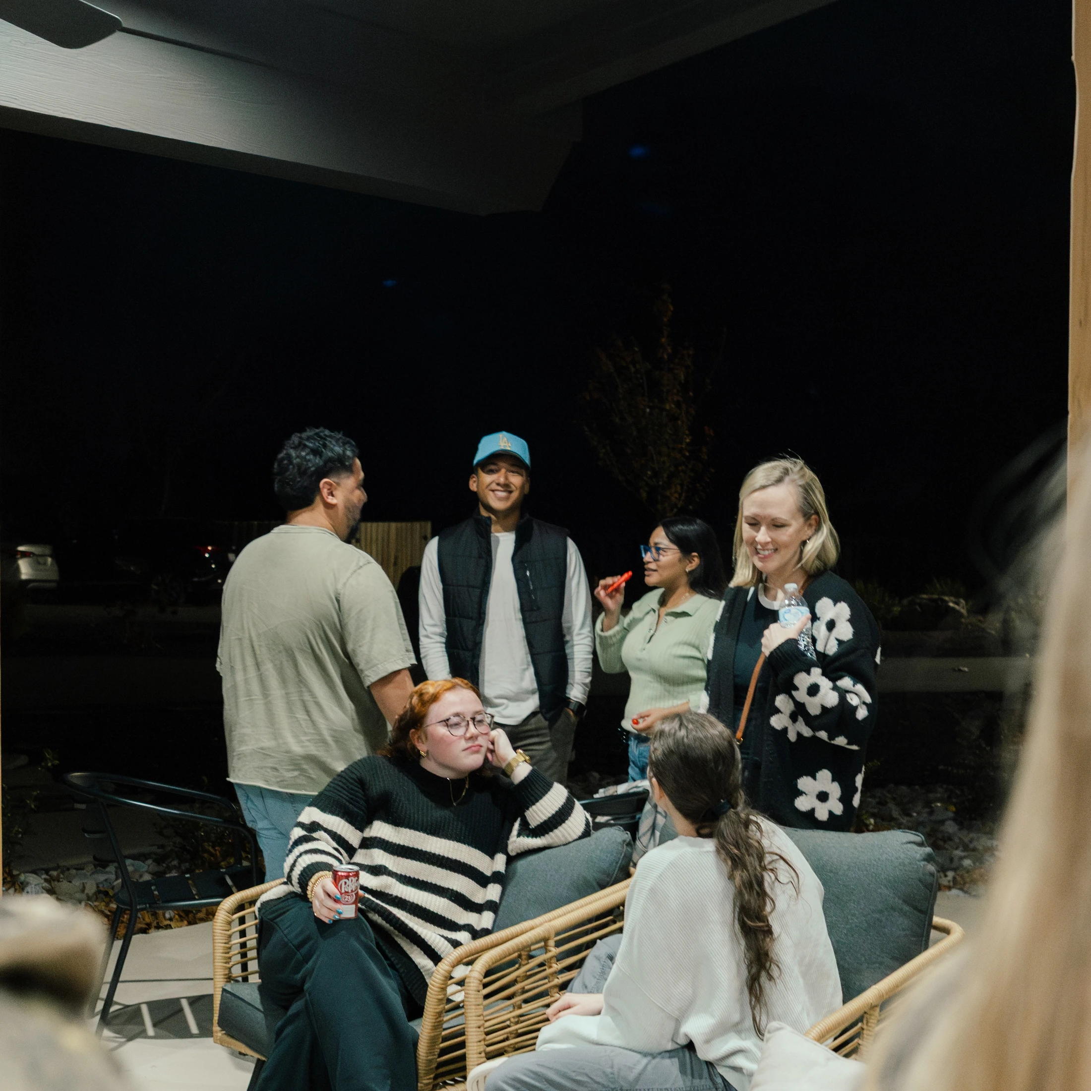 A diverse group of six young adults socializing on a covered patio at night. They are talking and smiling in a relaxed, friendly gathering.