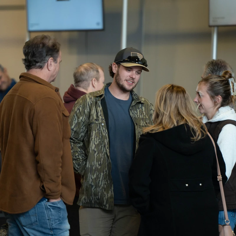 A group of people stand together indoors and talk. A young man in a camouflage jacket and baseball cap is the focus, smiling as he speaks with two women.
