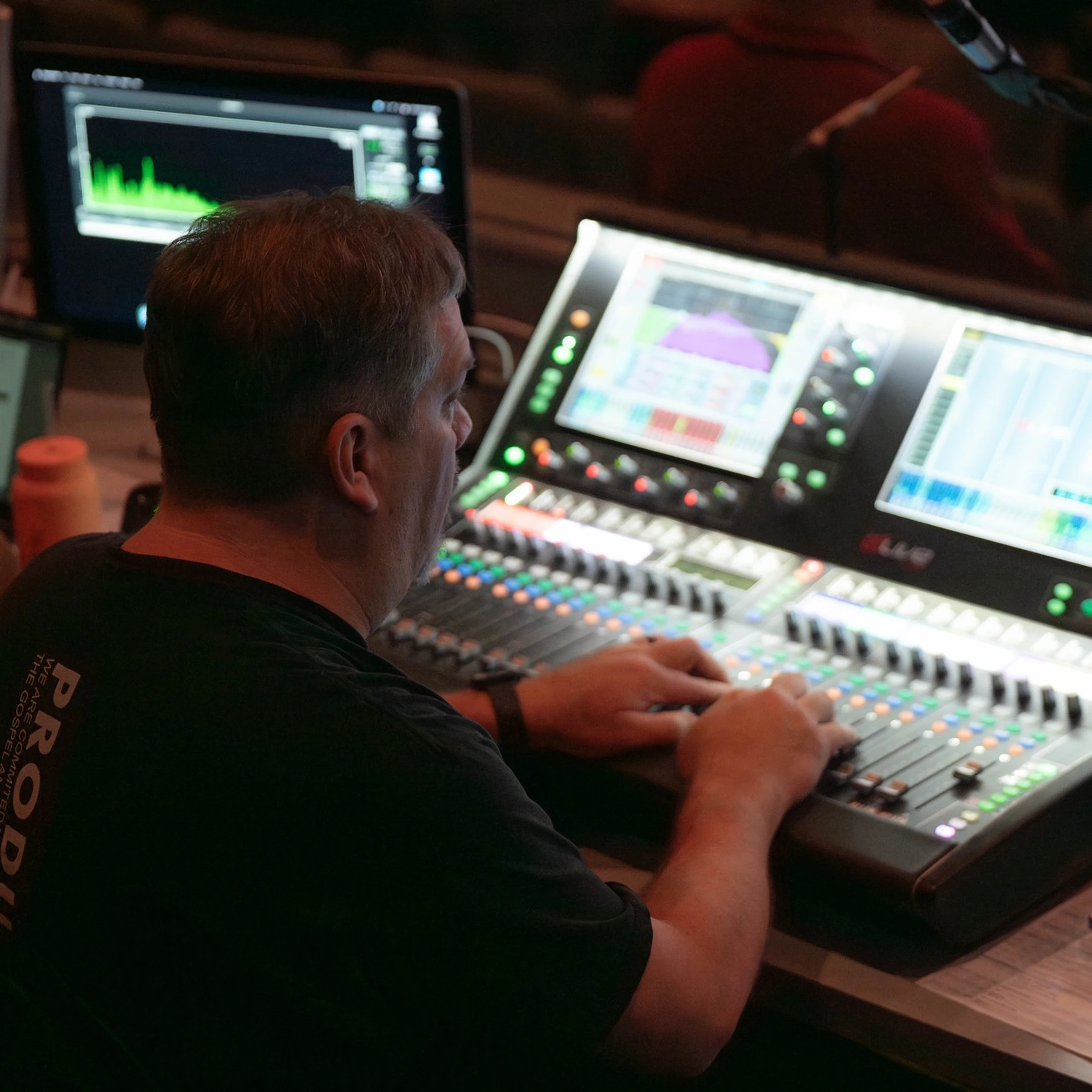 A sound engineer seen from behind, operating a large, illuminated audio mixing board with multiple screens displaying waveforms in a dimly lit room.