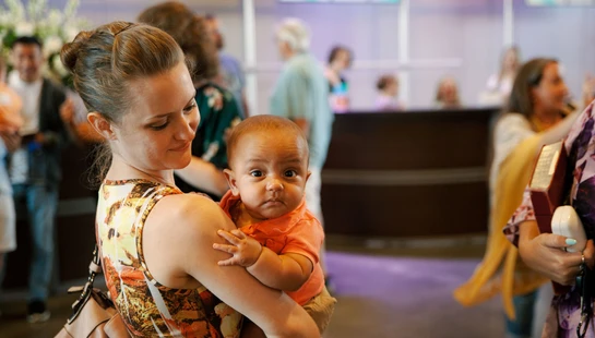 A woman with her hair in a bun holds a small baby in an orange shirt. The woman looks down at the baby, who is looking directly at the camera with wide eyes.