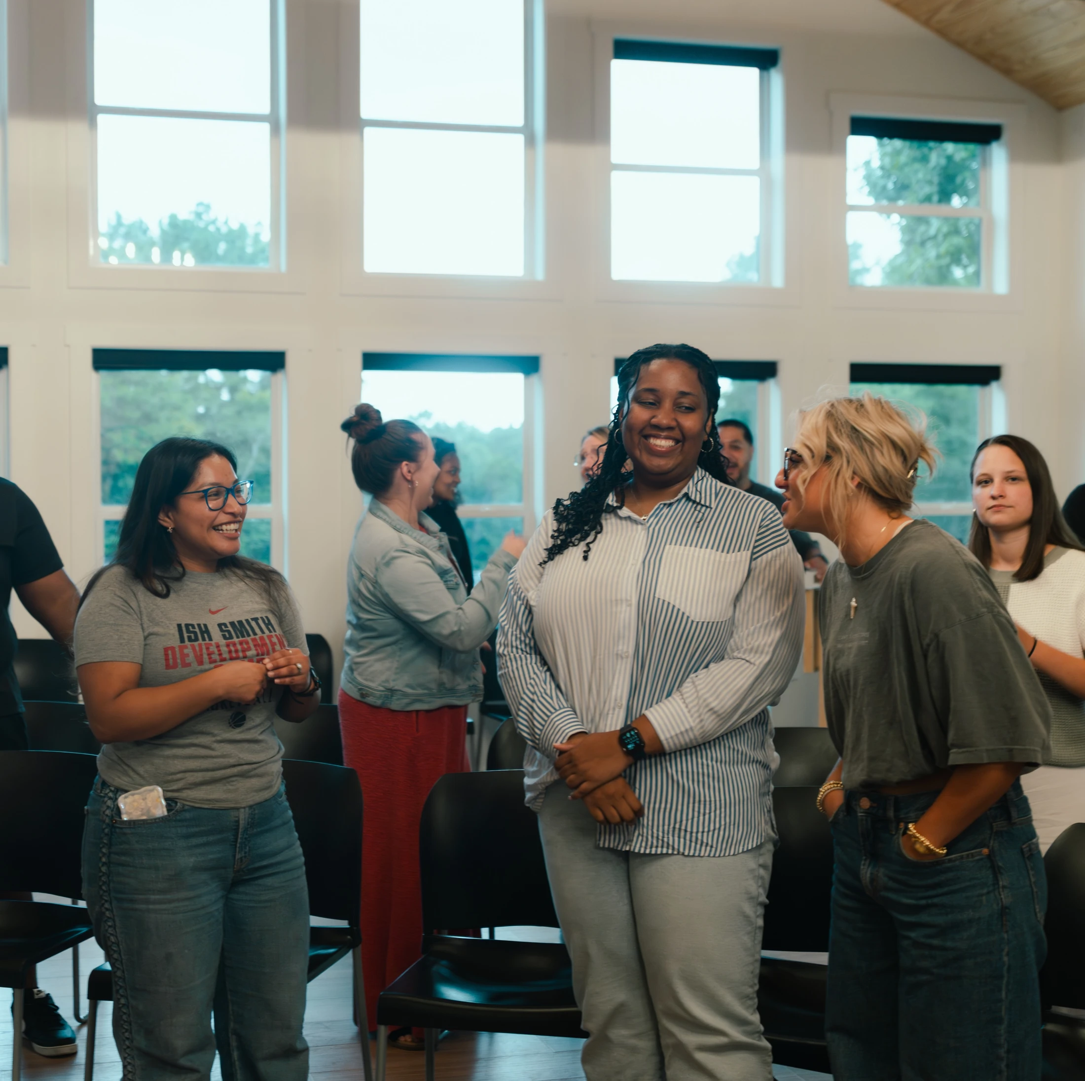 A group of diverse people stand and talk in a bright room with large windows. Three women in the foreground are smiling and engaged in conversation.