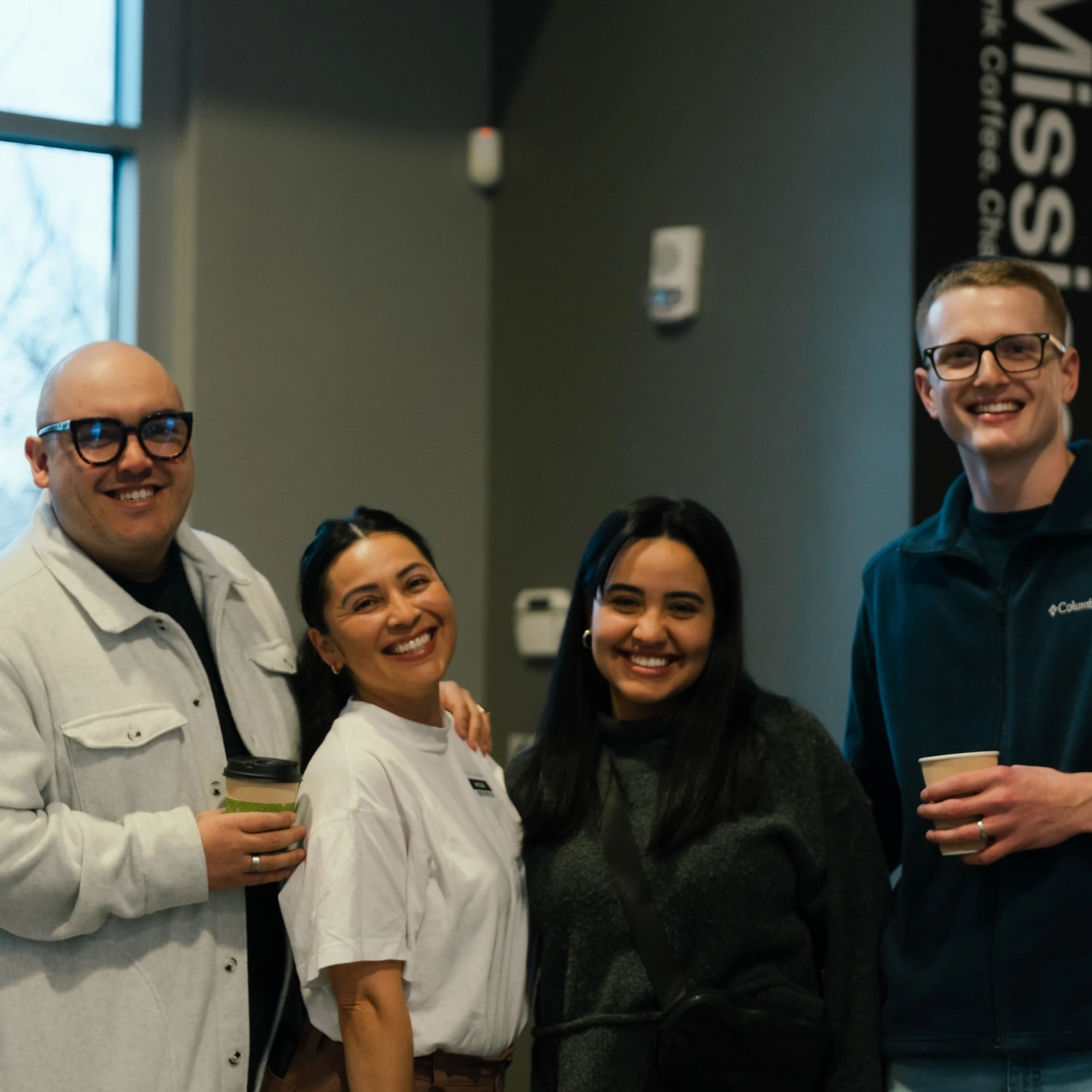 A group of four diverse people, two men and two women, smiling for the camera while standing together indoors. Two of them are holding coffee cups.
