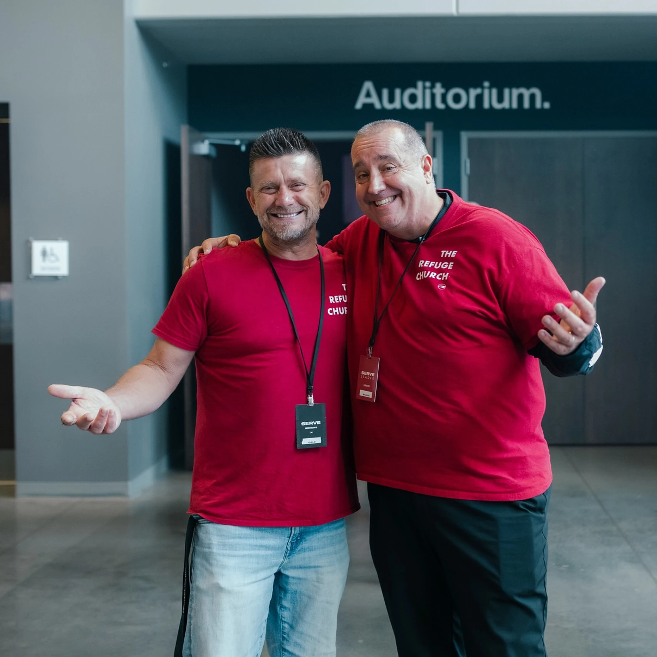Two smiling men in matching red t-shirts for 'The Refuge Church' stand arm-in-arm outside an auditorium, gesturing a warm welcome.