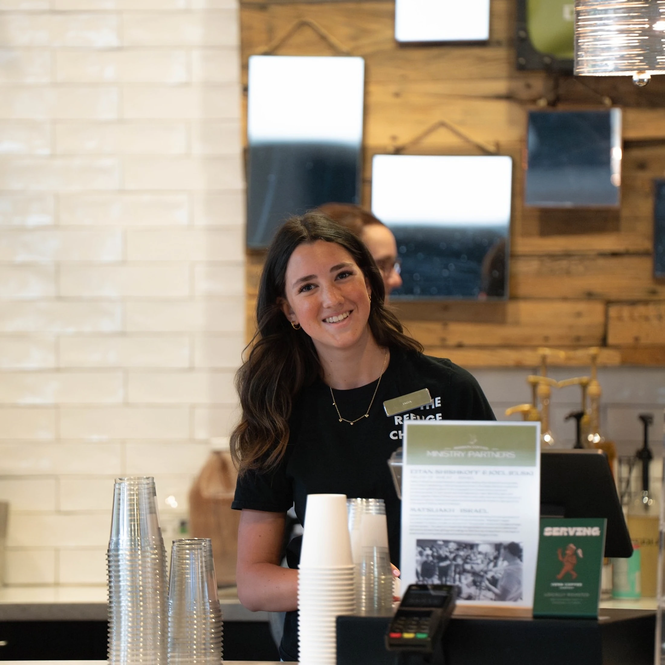 A young woman with long dark hair wearing a black shirt and a name tag smiles warmly while standing behind the counter of a modern cafe.