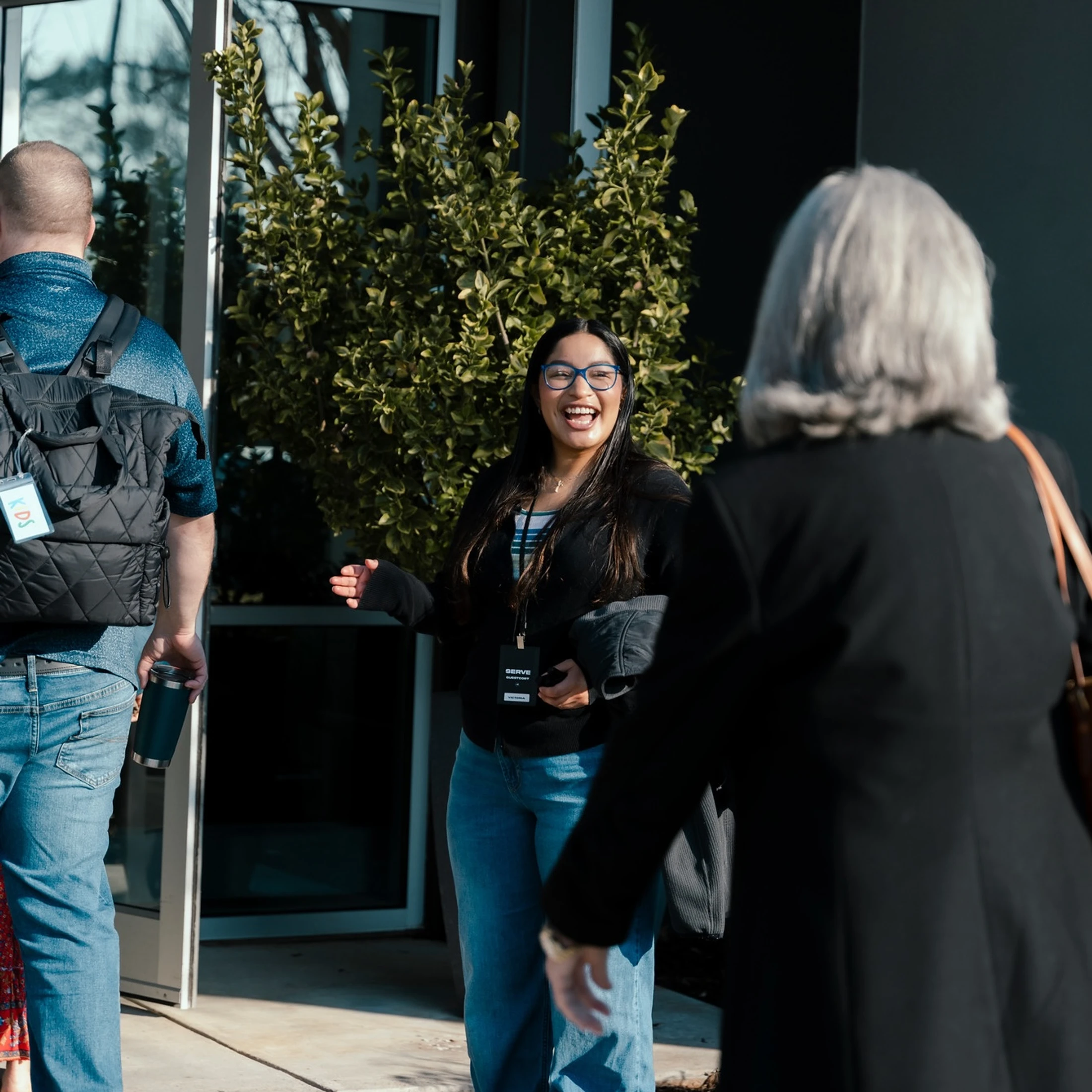 A young woman with glasses and a lanyard smiles warmly as she greets a man with a backpack and a woman with grey hair outside a building on a sunny day.