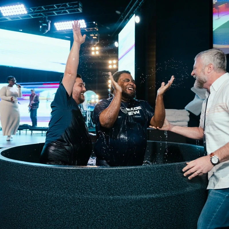 Two men celebrate in a baptismal pool on a church stage, splashing water, while a pastor smiles at them. A worship band performs in the background.