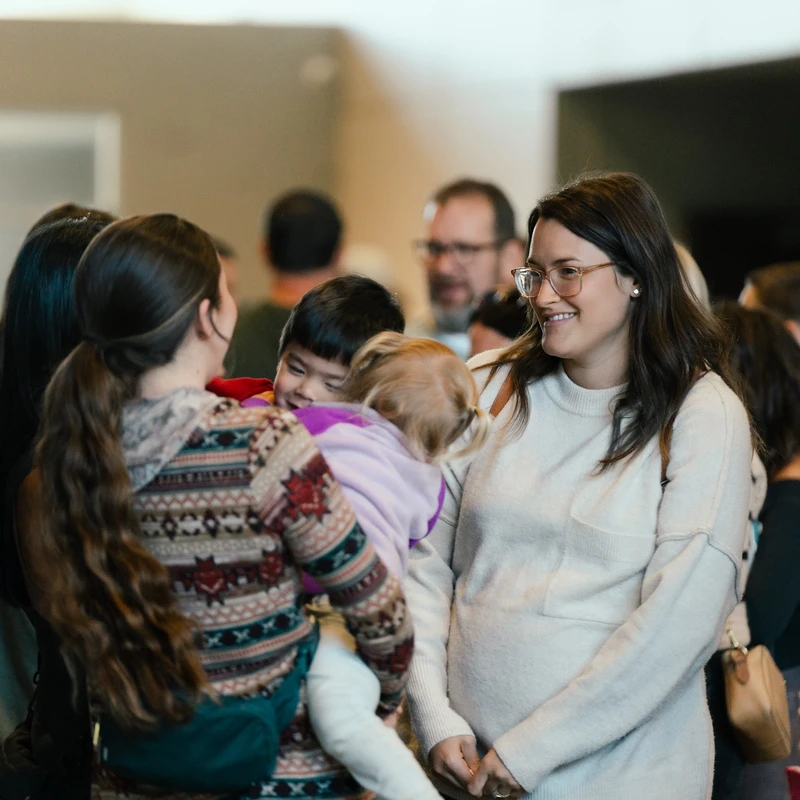 A group of people, including women and children, socializing indoors. A smiling woman in a cream sweater talks to another woman holding a small child.