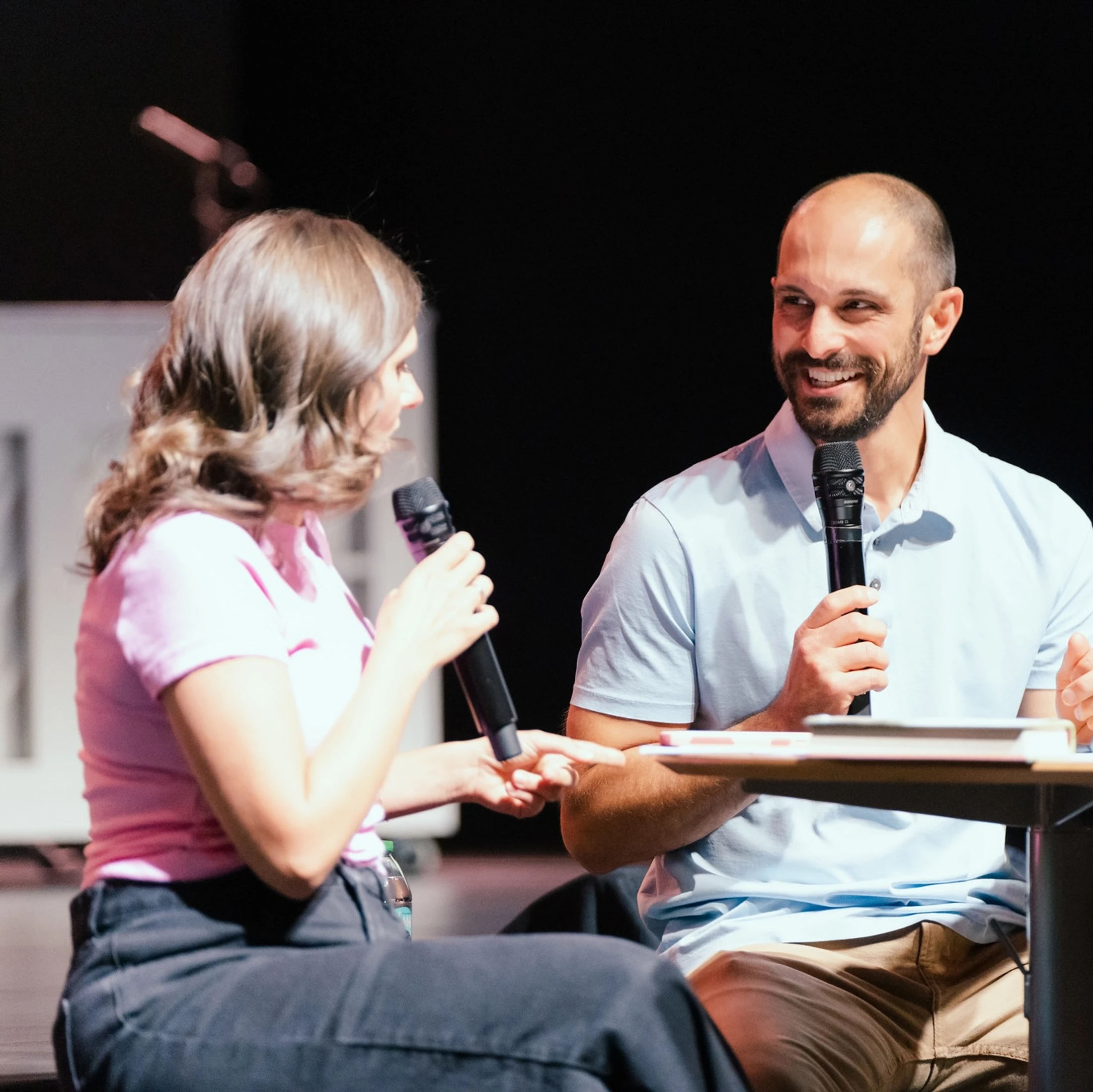 A smiling man with a microphone looks at a woman holding a microphone as they sit at a table on a stage during an event, engaged in conversation.