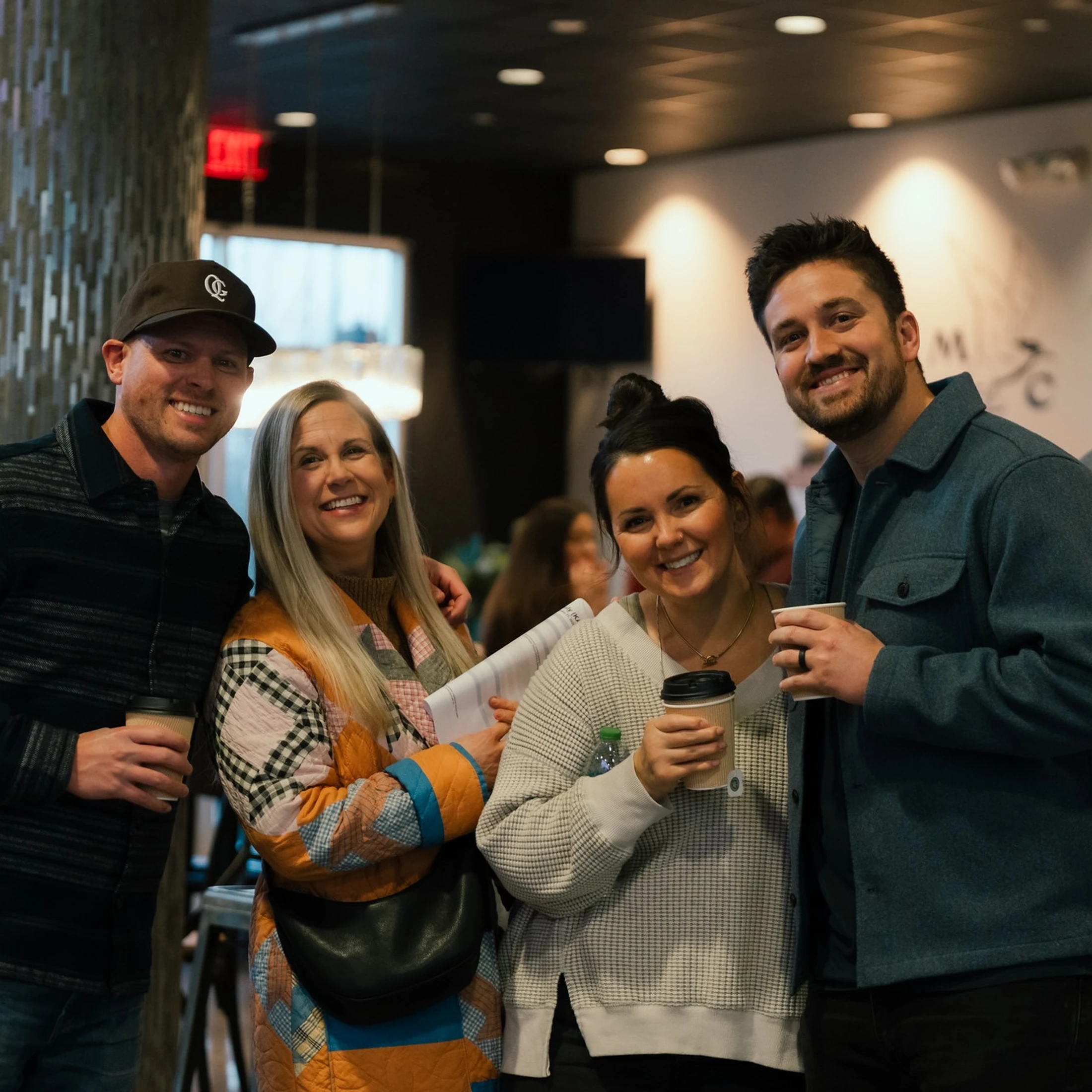 Four smiling friends, two men and two women, stand close together indoors, holding coffee cups and posing for a picture in a warmly lit room.
