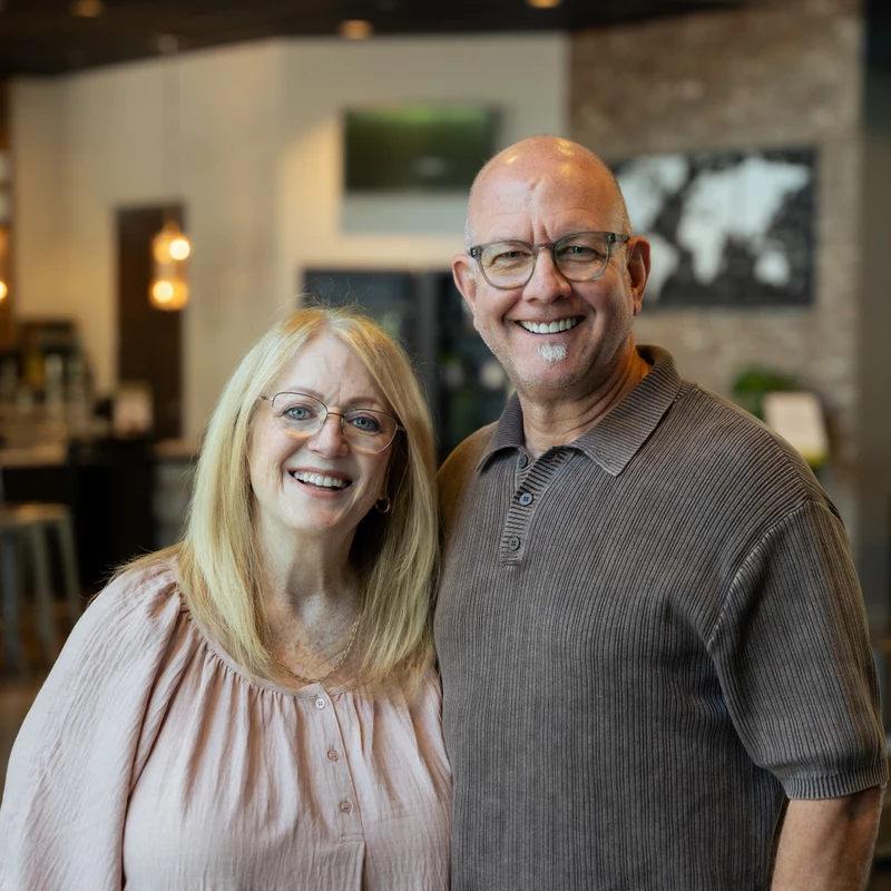 A happy middle-aged couple, a woman with blonde hair and a man with glasses, smile while posing for a photo together in a modern indoor setting.