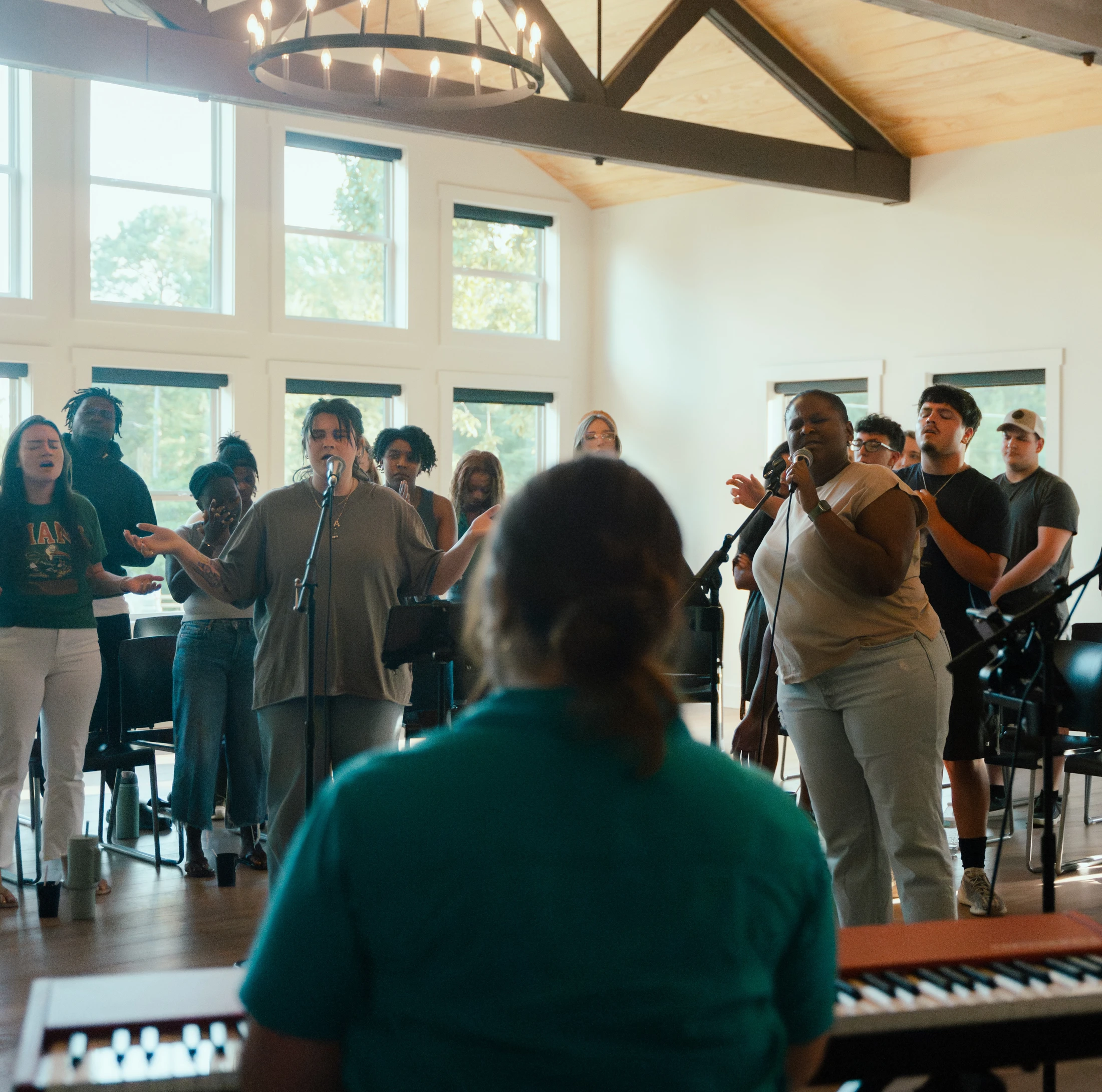 From behind a keyboard player, a diverse group of people sing together in a bright, spacious room. Two women at the front lead with microphones.