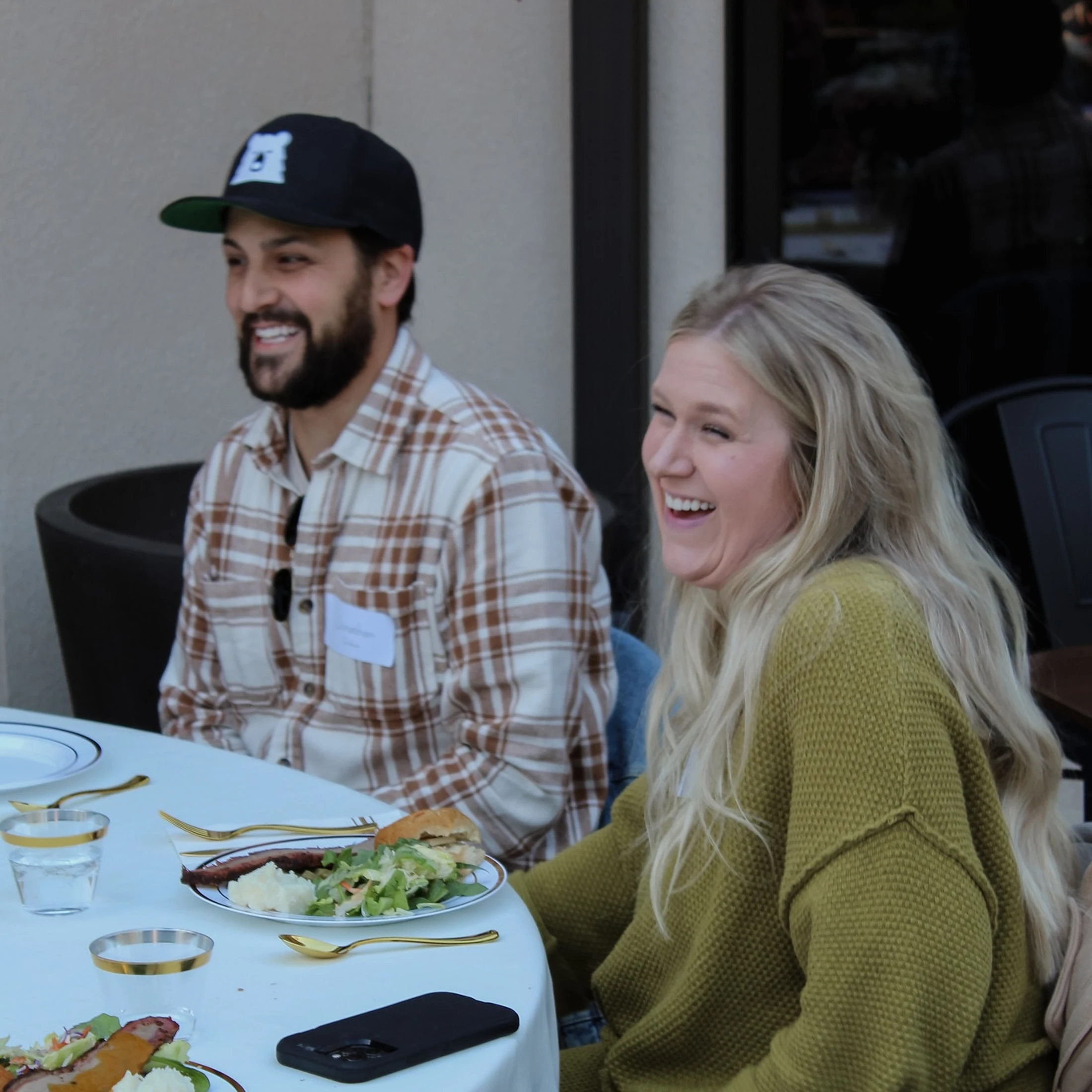 A man in a plaid shirt and cap smiles while a woman with long blonde hair in a green sweater laughs. They are sitting at a dining table with plates of food.