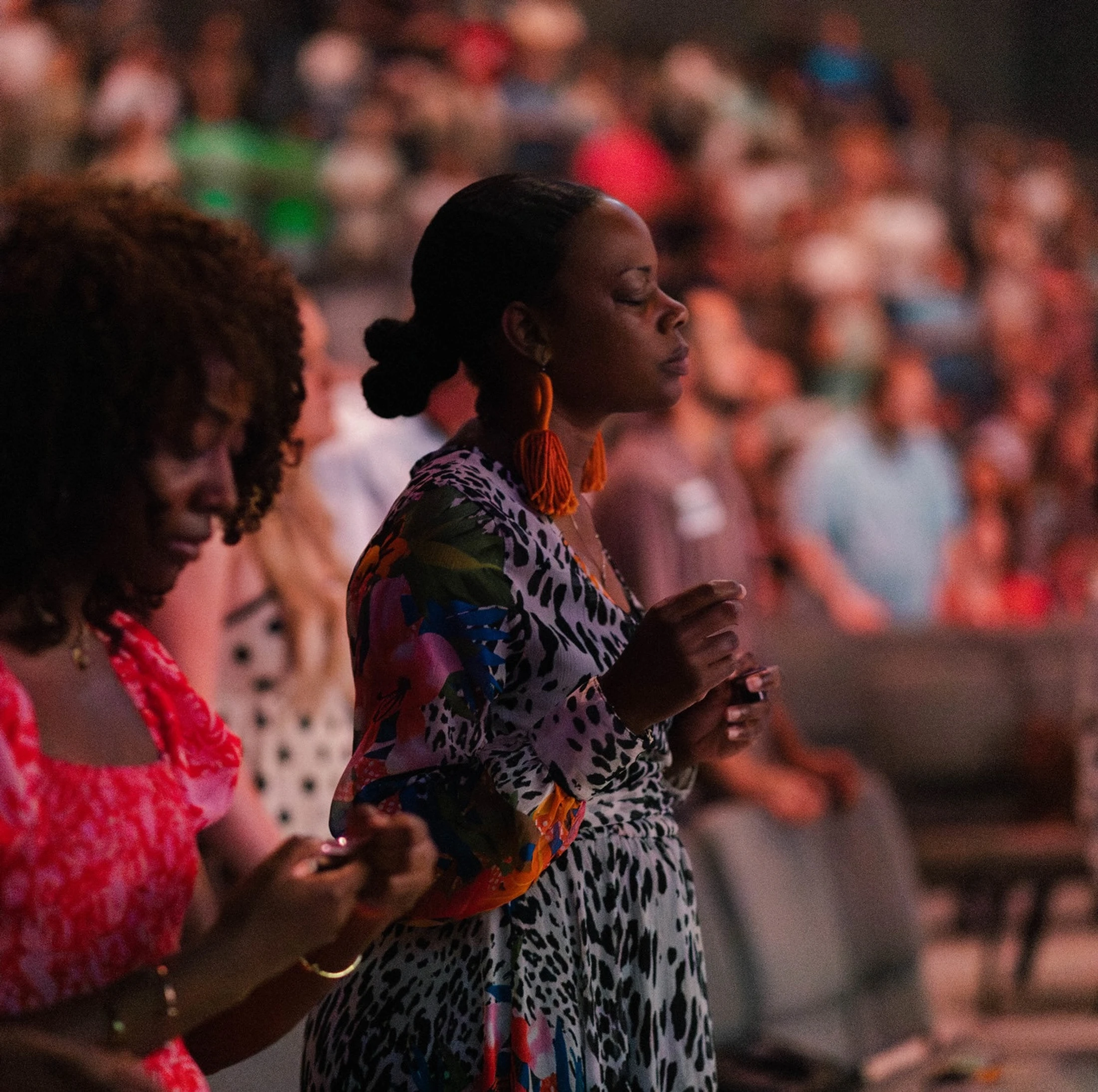 A Black woman with her eyes closed stands in prayer amidst a large, blurred crowd. She wears a colorful patterned dress and large orange tassel earrings.