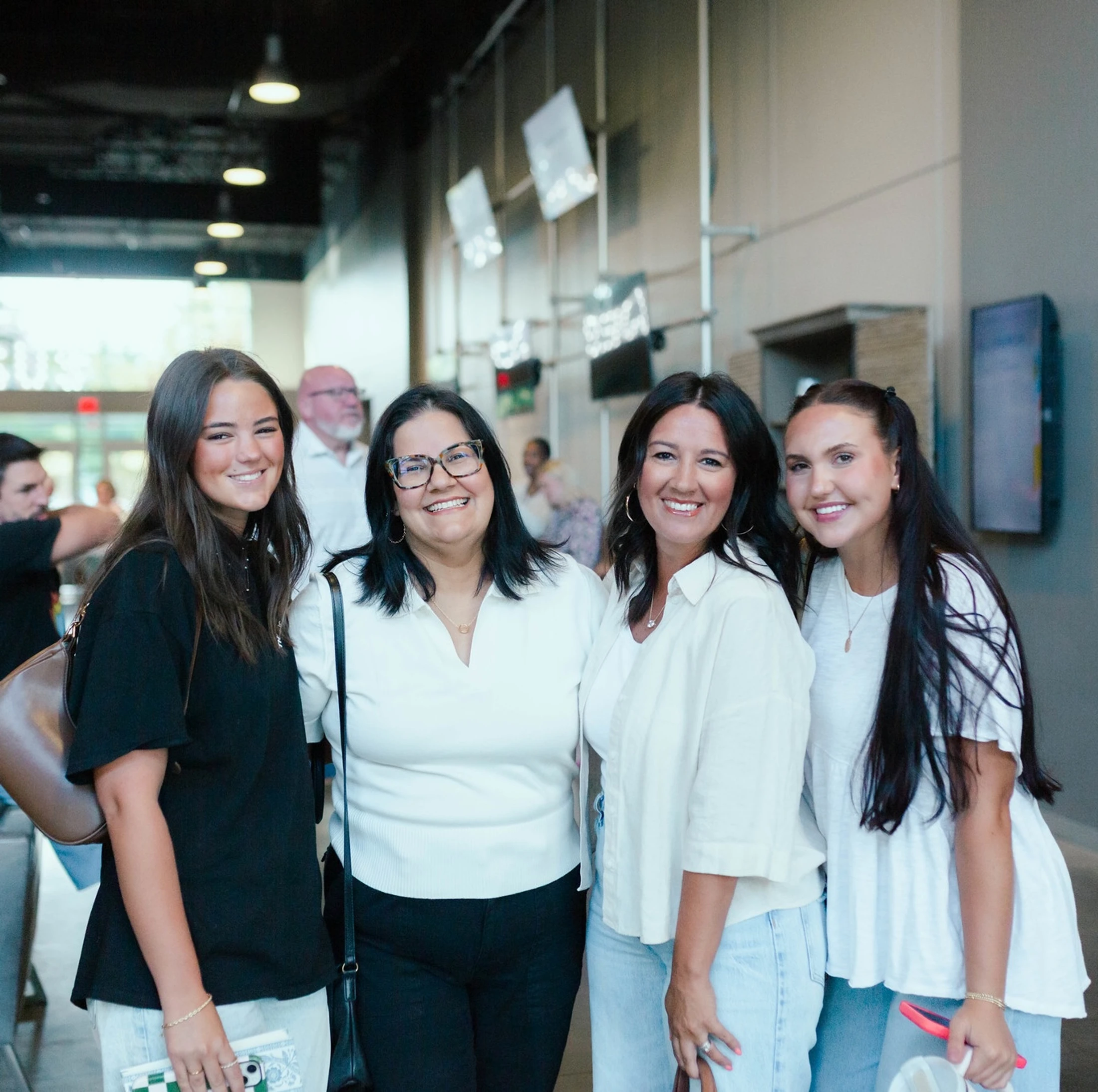 A group of four women stand closely together, smiling for the camera inside a modern building lobby during an event.