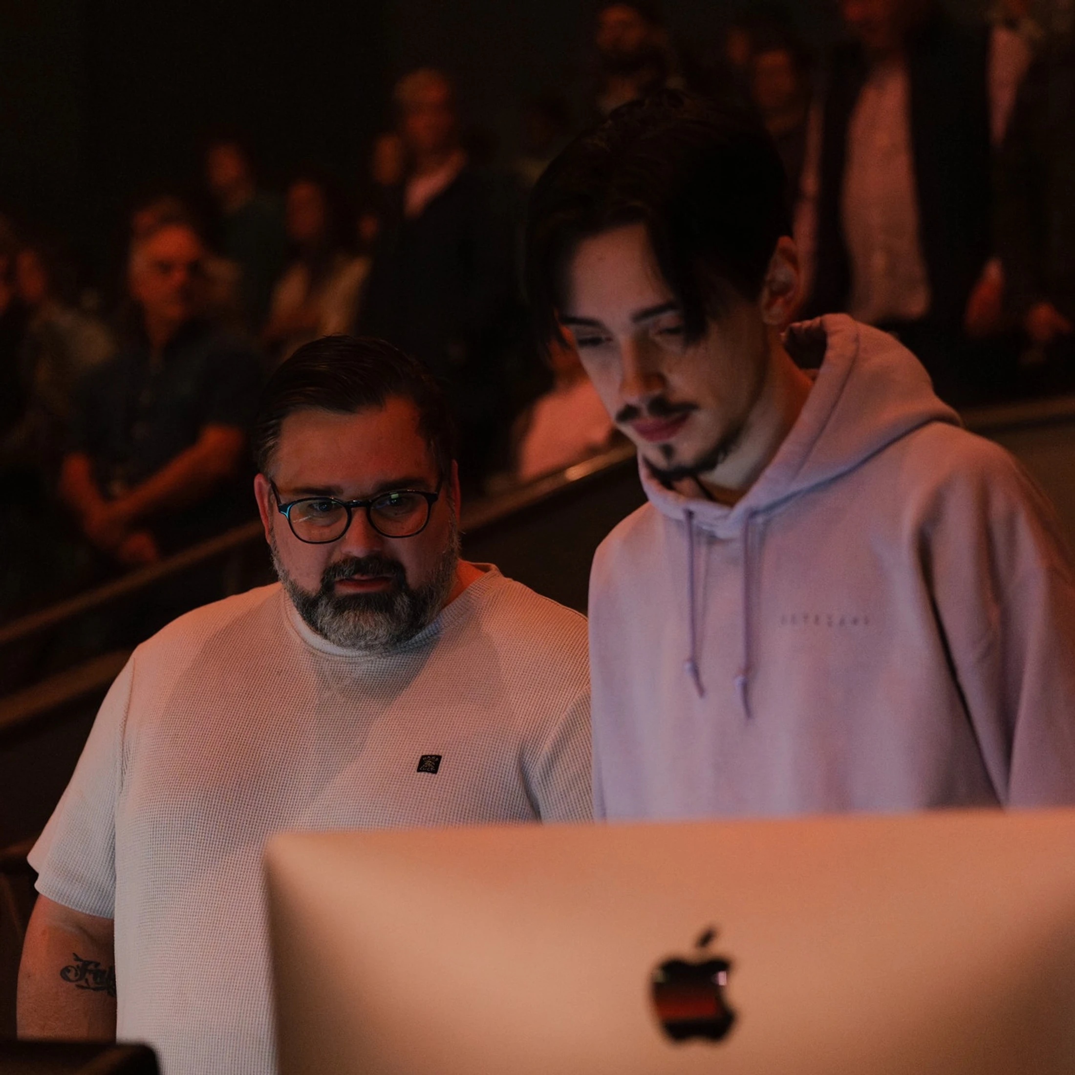 An older man with a beard and glasses stands next to a younger man in a hoodie. They are both looking down at an Apple computer screen in a dimly lit room.