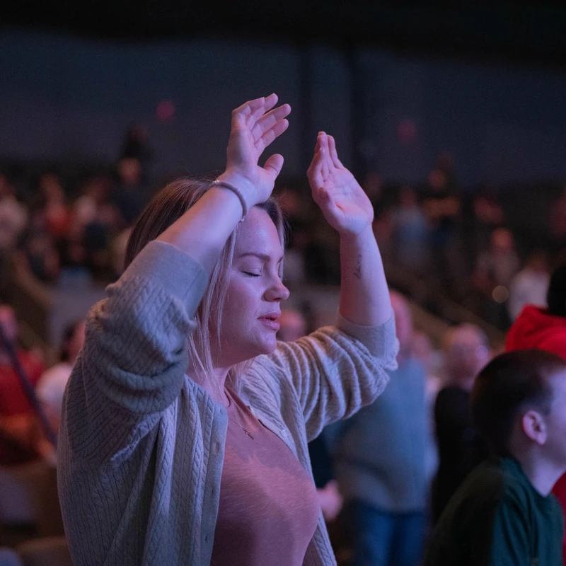 A woman with blonde hair and a tan sweater lifts her hands with her eyes closed in an act of worship, surrounded by a congregation in a dimly lit room.