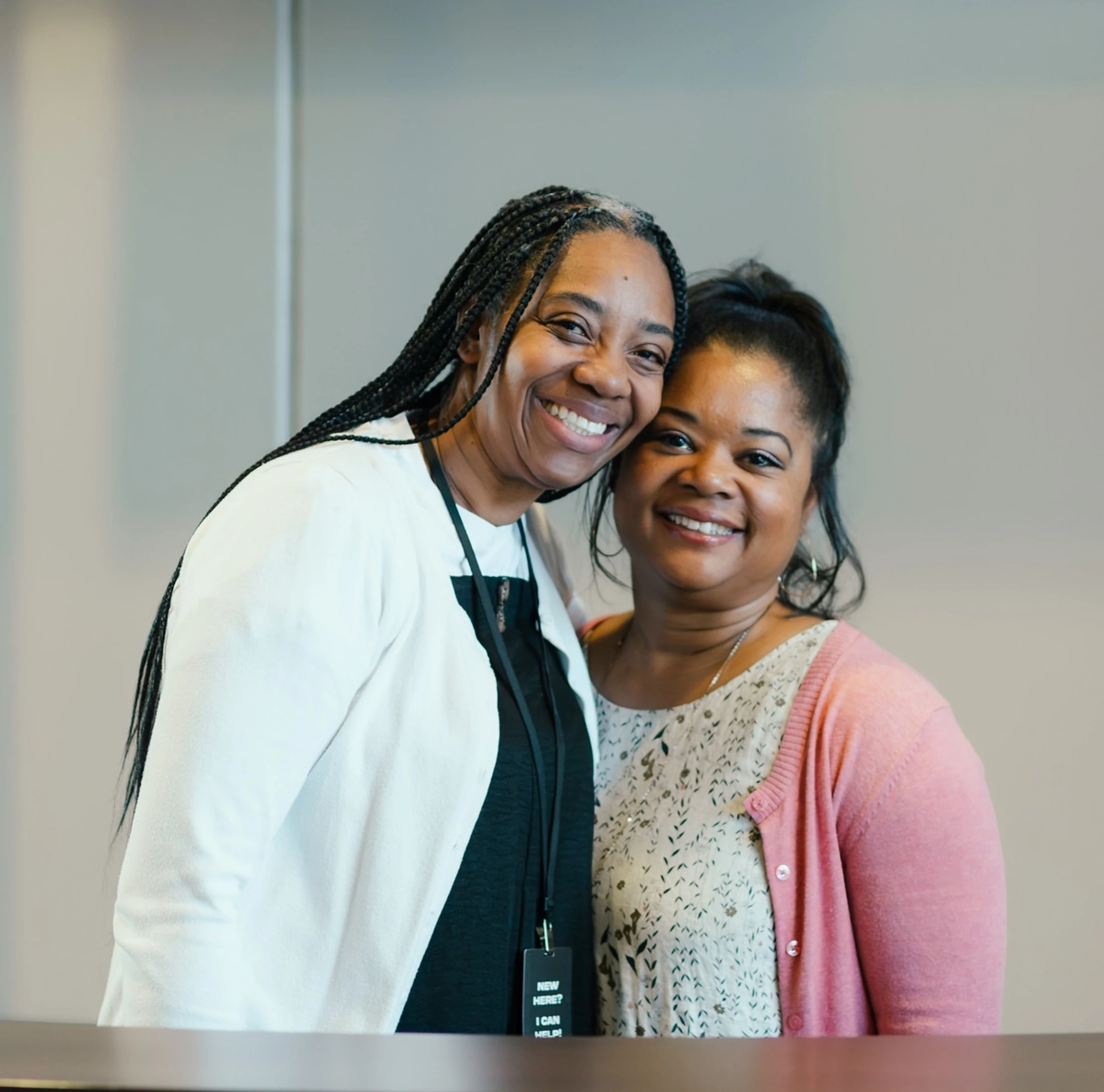 A portrait of two smiling Black women standing close together. The woman on the left has long braids and wears a lanyard that reads 'I CAN HELP'.