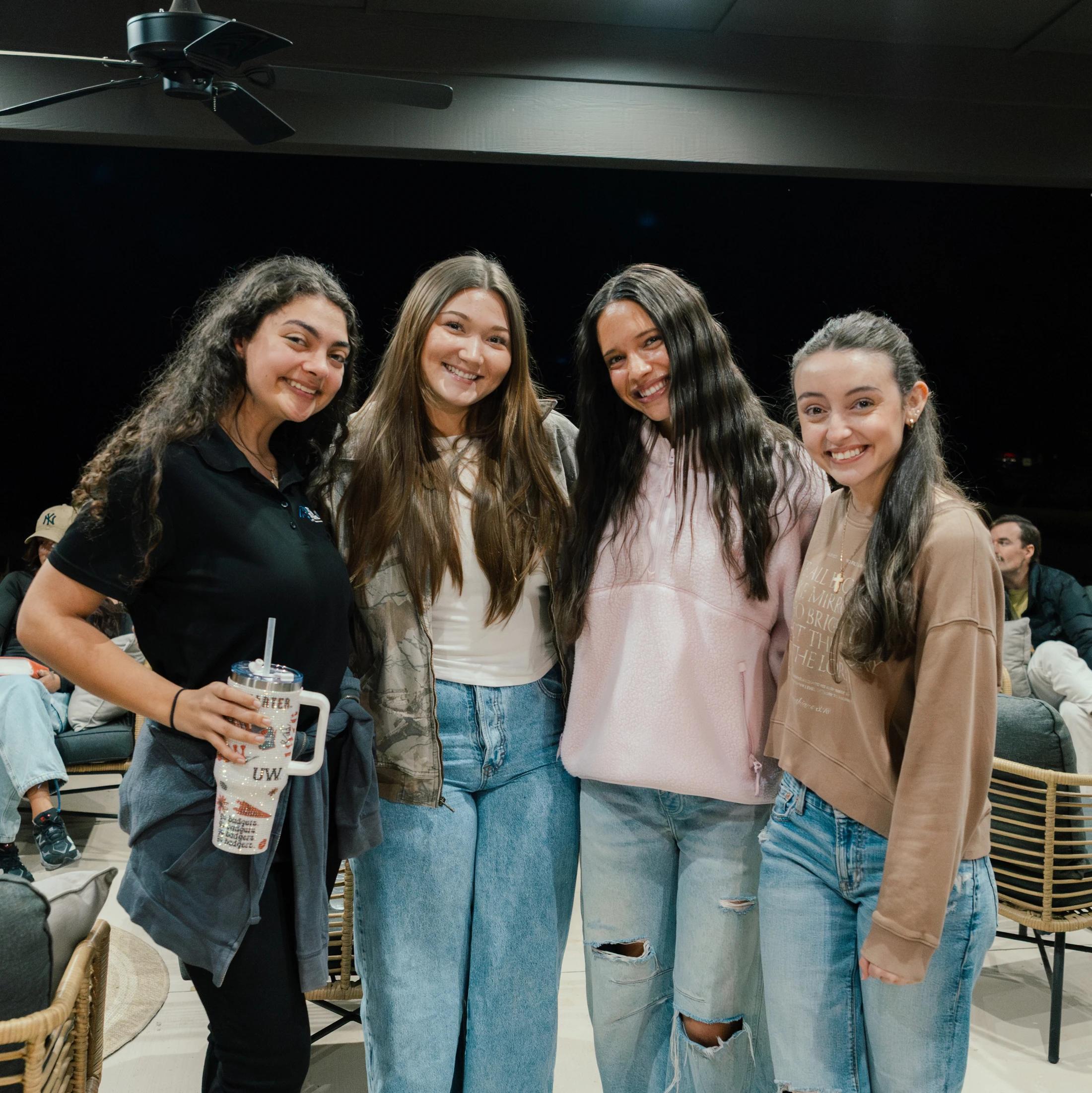 A group of four young women stand close together, smiling for the camera. They are dressed in casual jeans and tops in a modern, relaxed indoor setting.