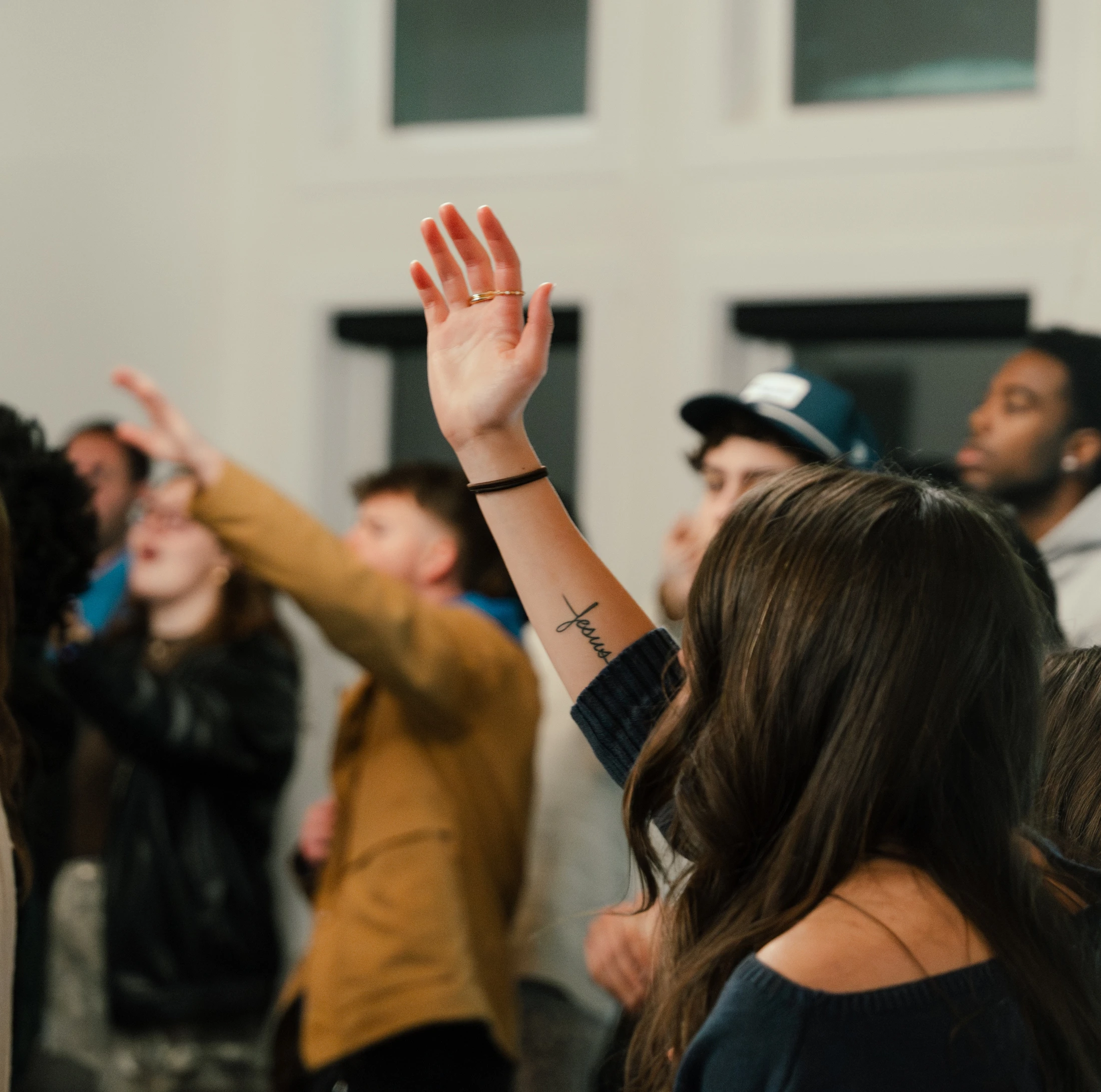 A woman seen from behind raises her hand among a group of people. Her forearm has a tattoo of the word 'Jesus' in cursive script.