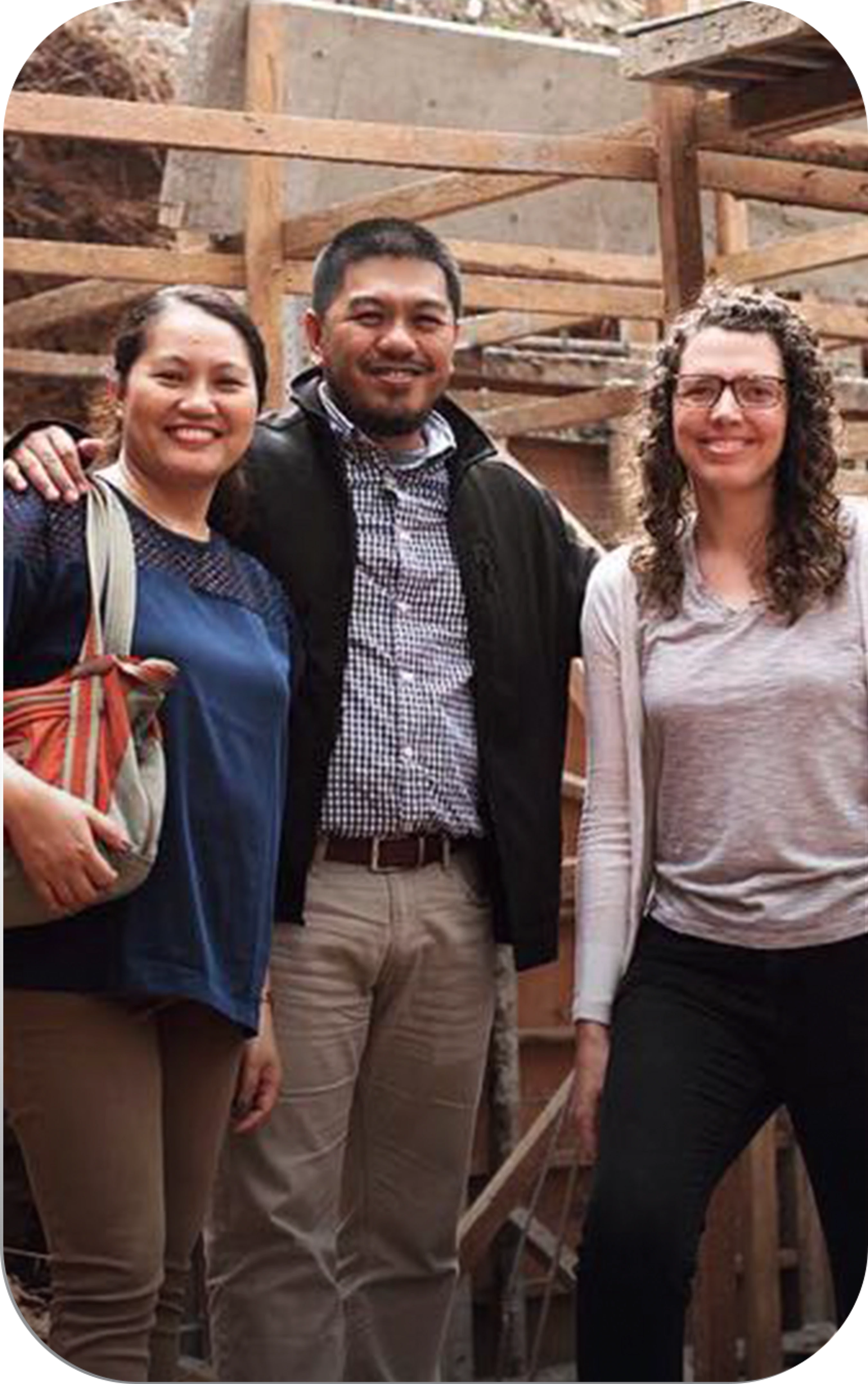 A man stands between two women, all smiling at the camera. They are outdoors in front of a building under construction with wooden framing visible.