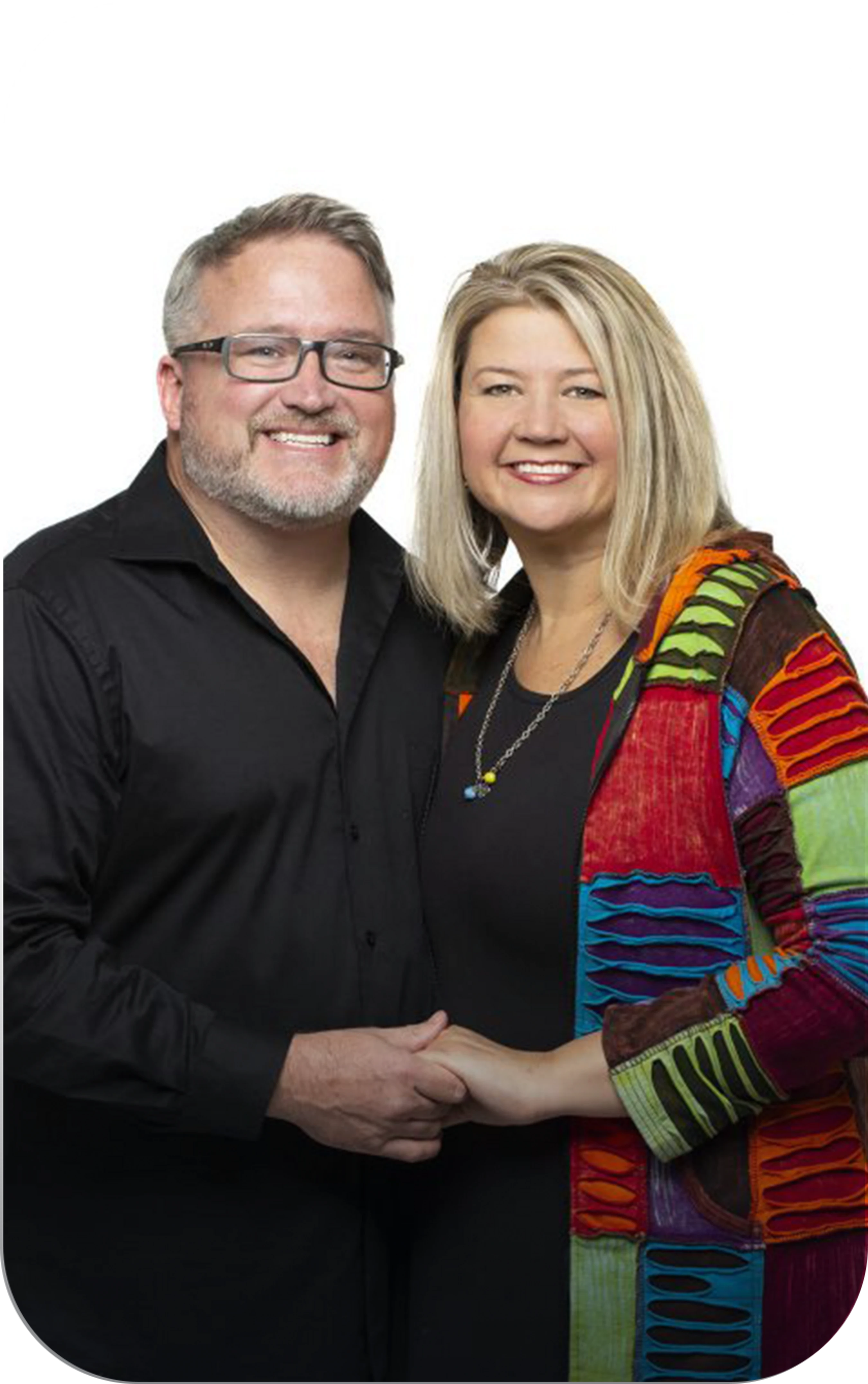 A smiling man and woman pose together against a white background. The man wears a black shirt and glasses, and the woman wears a colorful patchwork jacket.