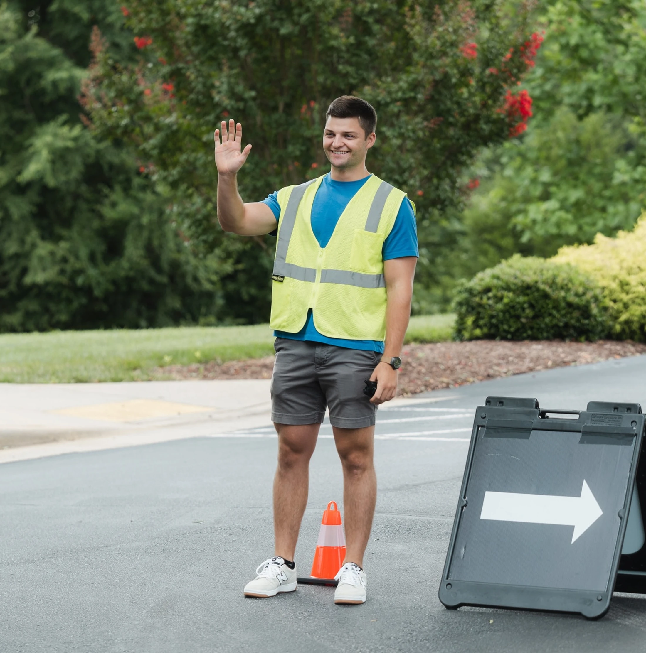 A smiling young man in a yellow safety vest waves while standing in a parking lot next to a cone and a directional sign with an arrow.