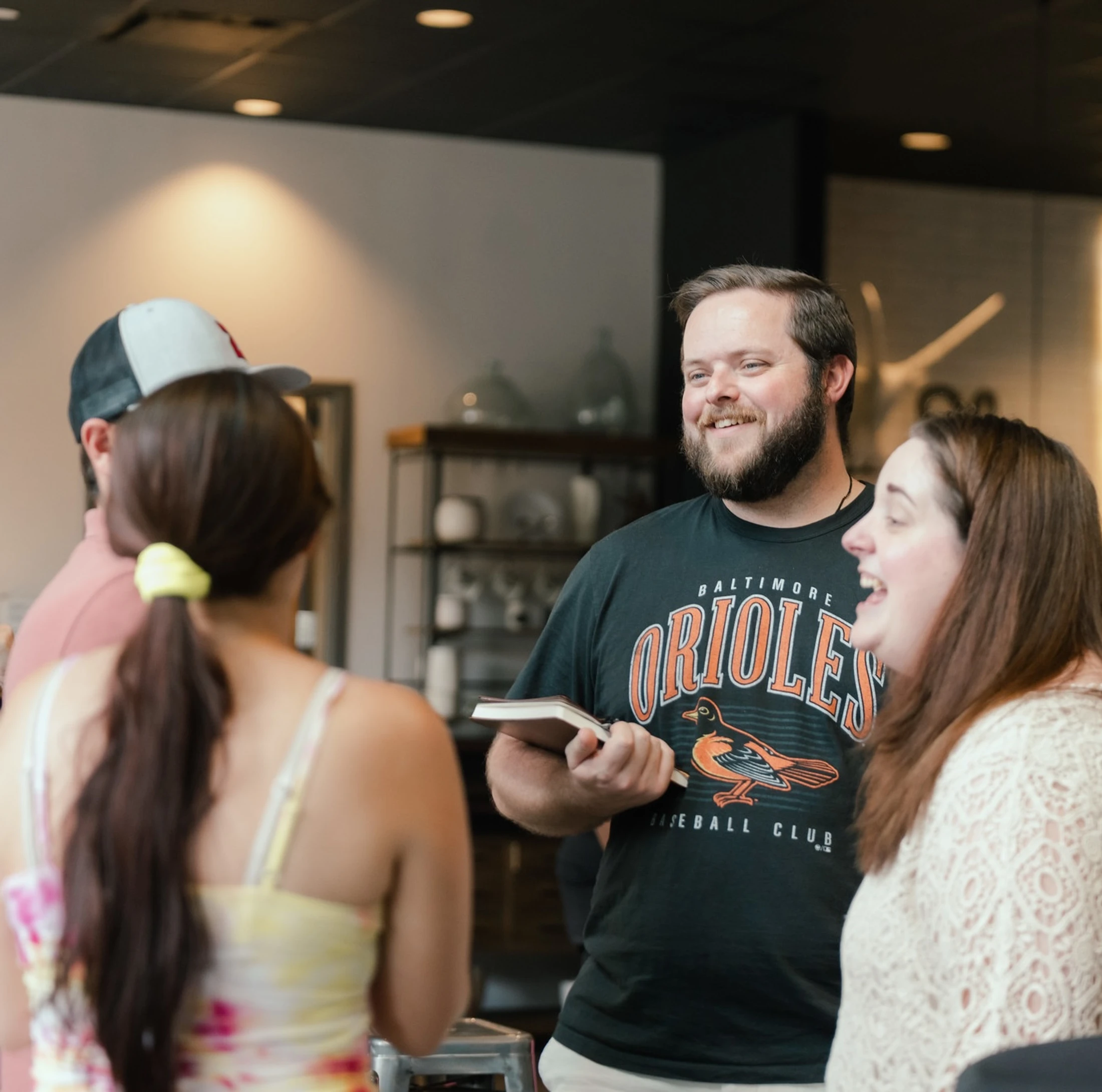 A group of people talking. A smiling man with a beard, wearing an Orioles t-shirt, holds a book and looks at a laughing woman next to him.