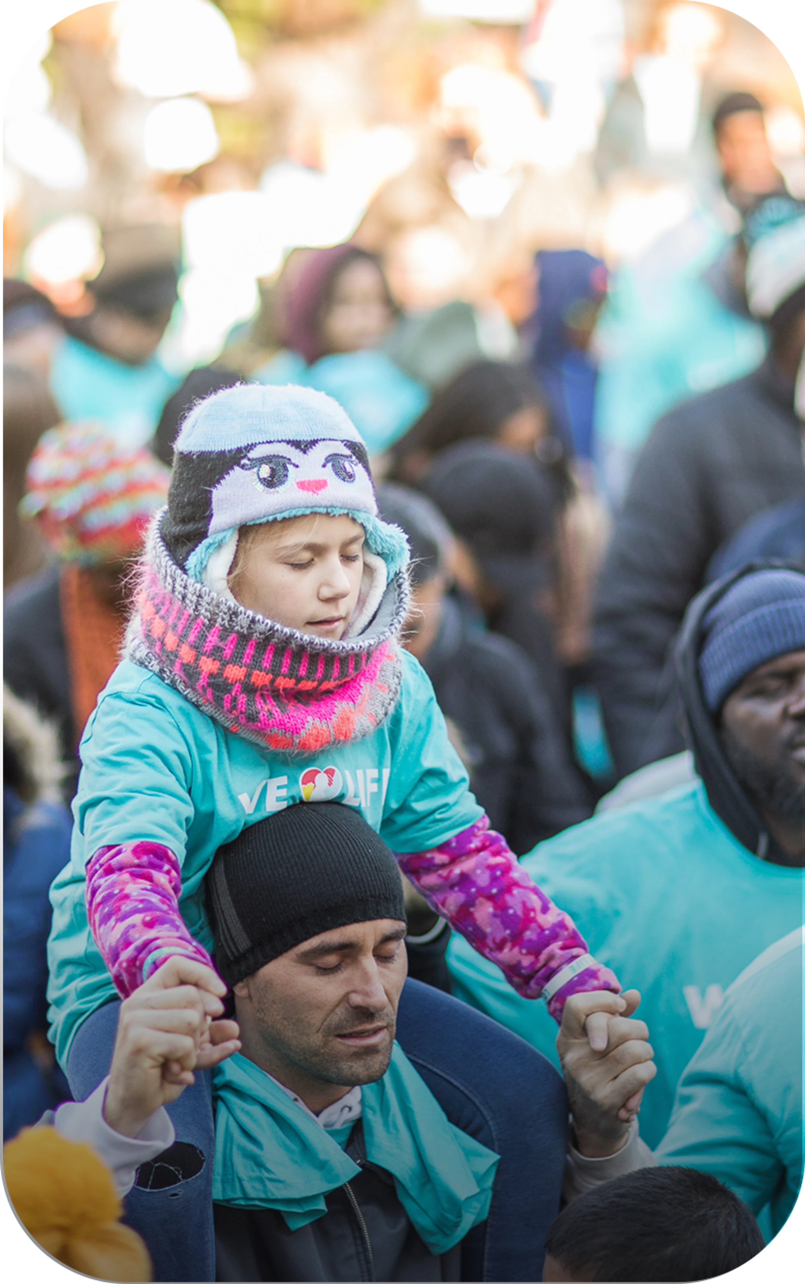 A young girl wearing a winter hat rides on a man's shoulders in a large crowd. Both have their eyes closed in a moment of quiet reflection during an event.