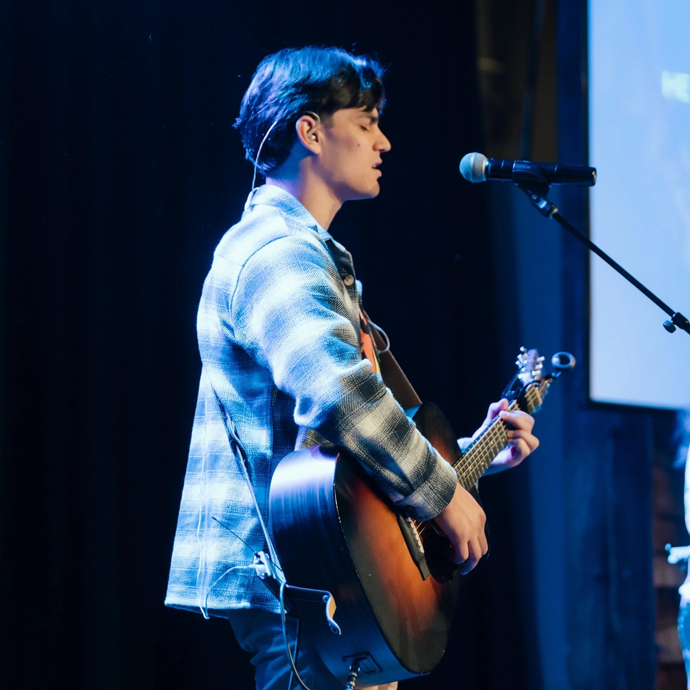 A young man in a plaid shirt plays an acoustic guitar and sings into a microphone on a stage bathed in cool blue light.